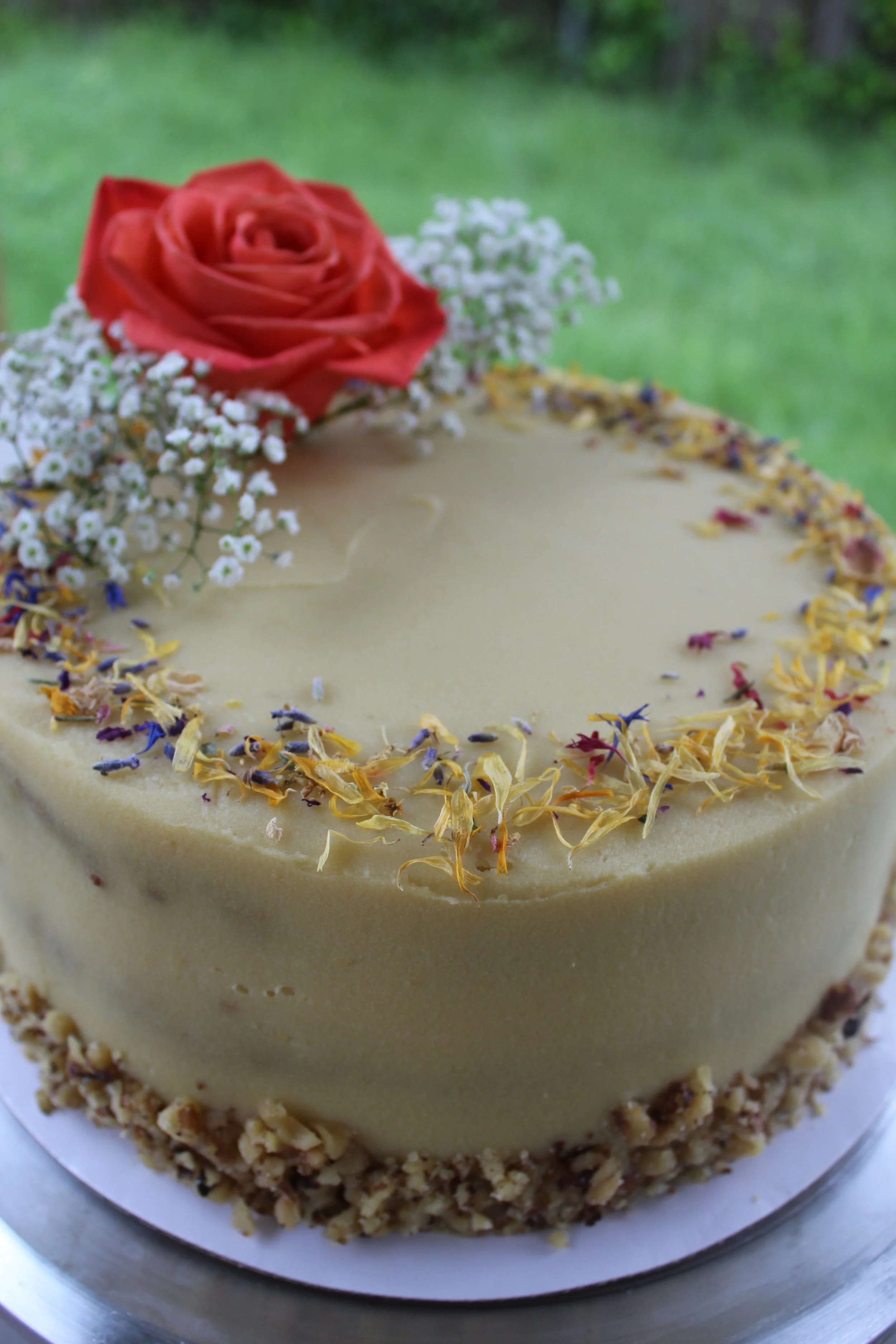 A round cake with white frosting, topped with a pink rose and white baby's breath flowers, and decorated with dried flower petals around the top edge, placed outdoors with a grassy background.