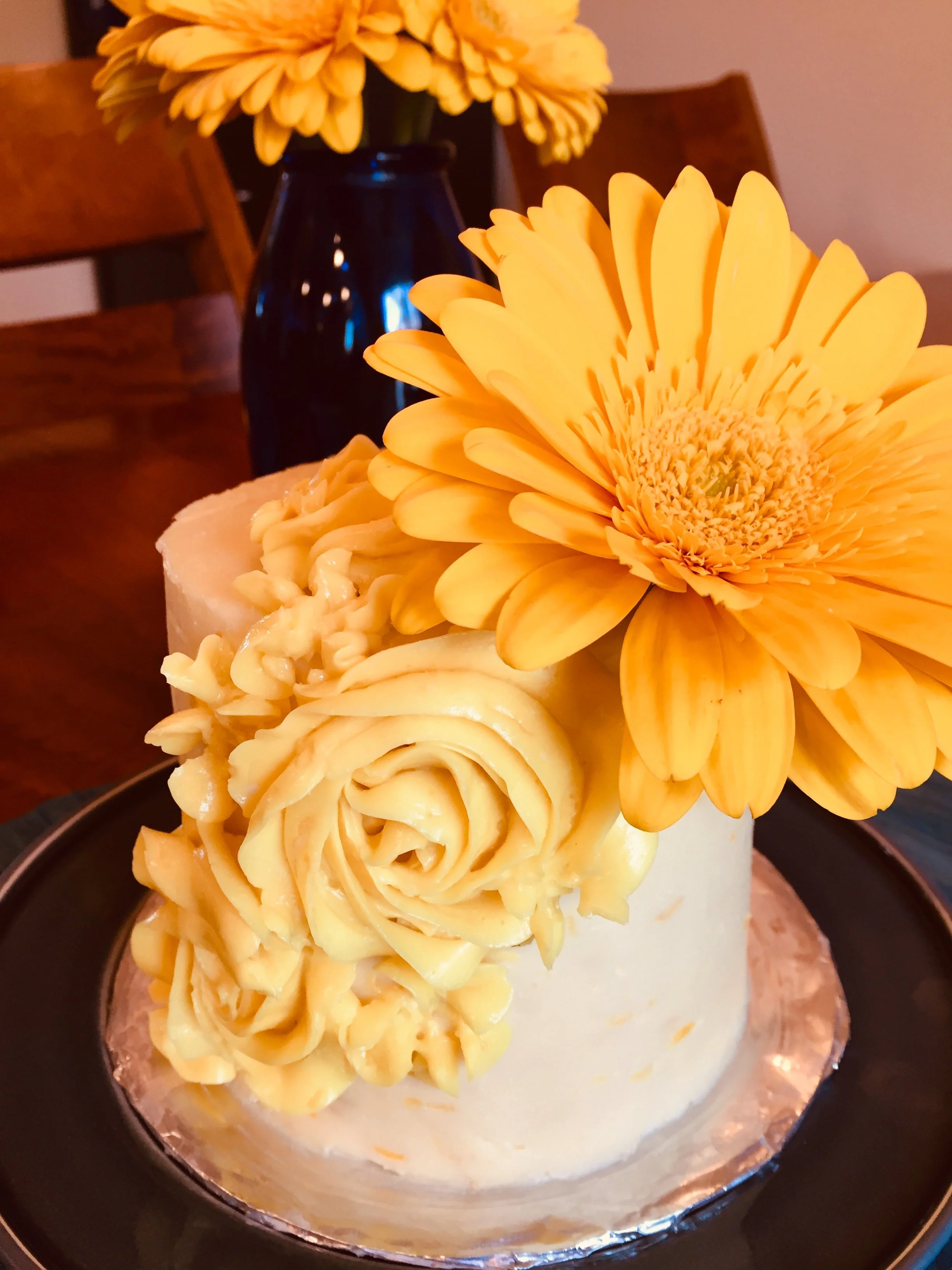 A cake decorated with large yellow daisies and roses, placed on a black plate with a gold-colored base.