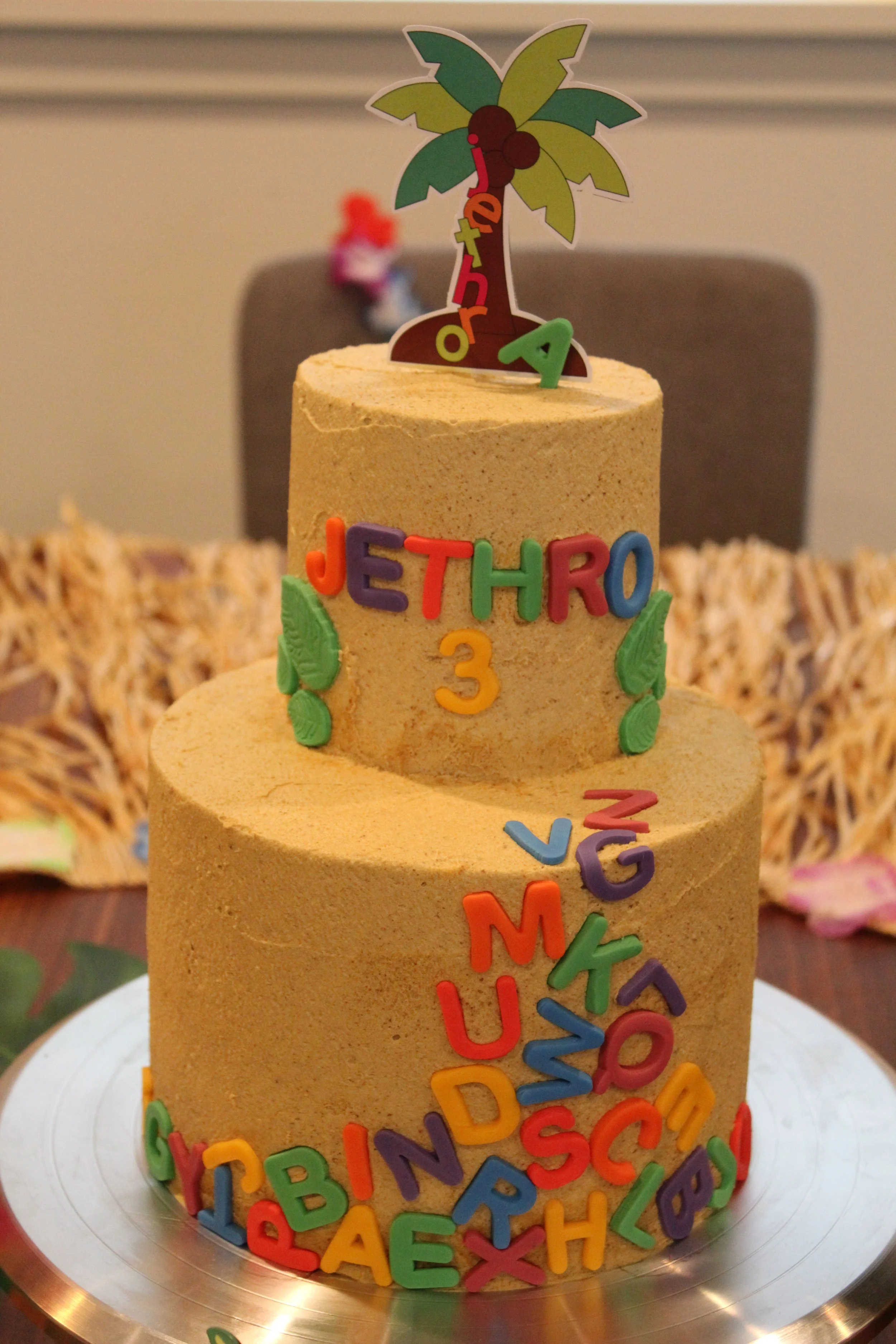 Two-tiered brown birthday cake decorated with colorful foam letter confetti, with the name JETHRO and the number 3 on it. The cake topper is a paper palm tree with the word 'Aloha' hanging from it.