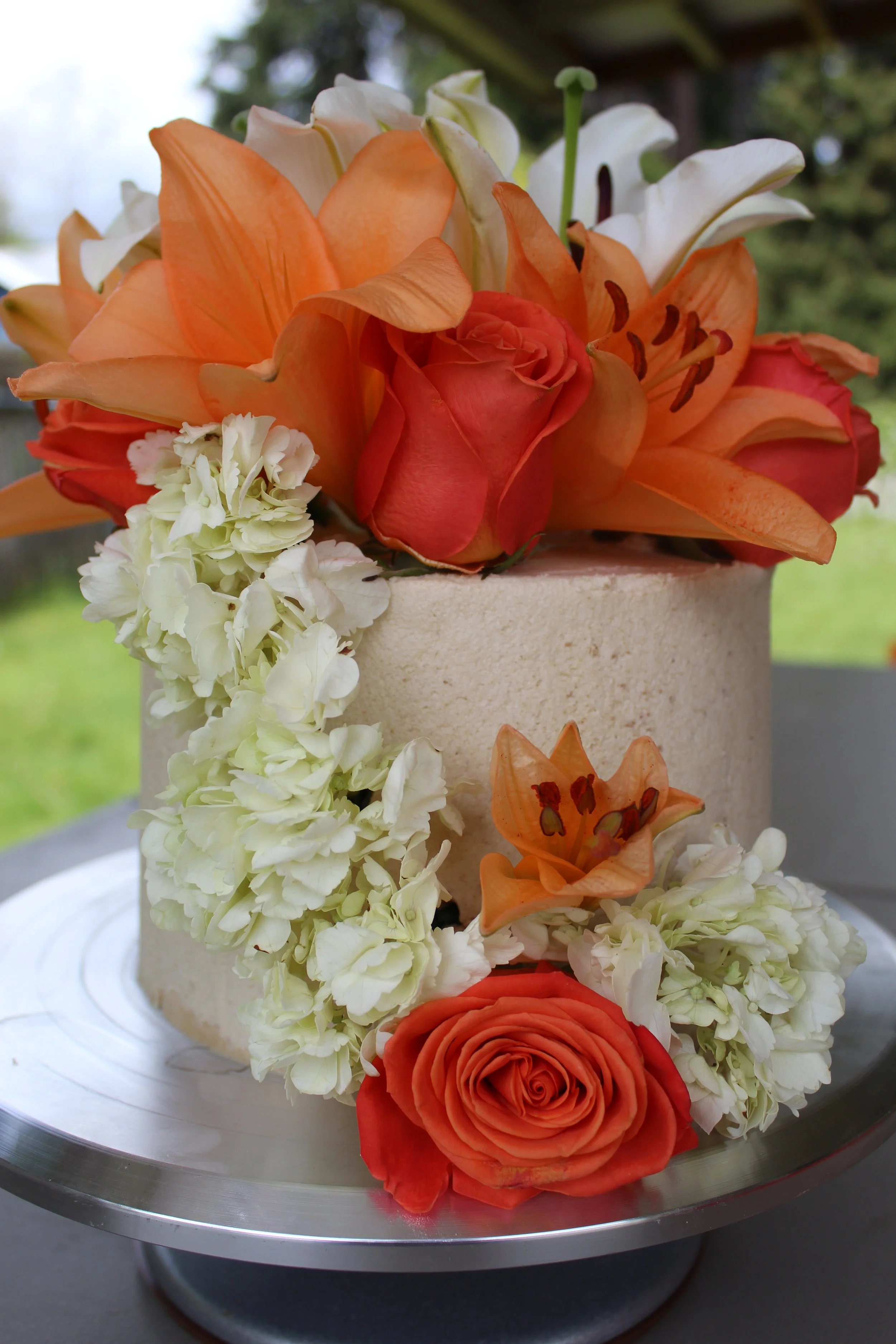 A decorated cake with orange and white flowers, including roses, lilies, and hydrangeas, on a silver cake stand outdoors.