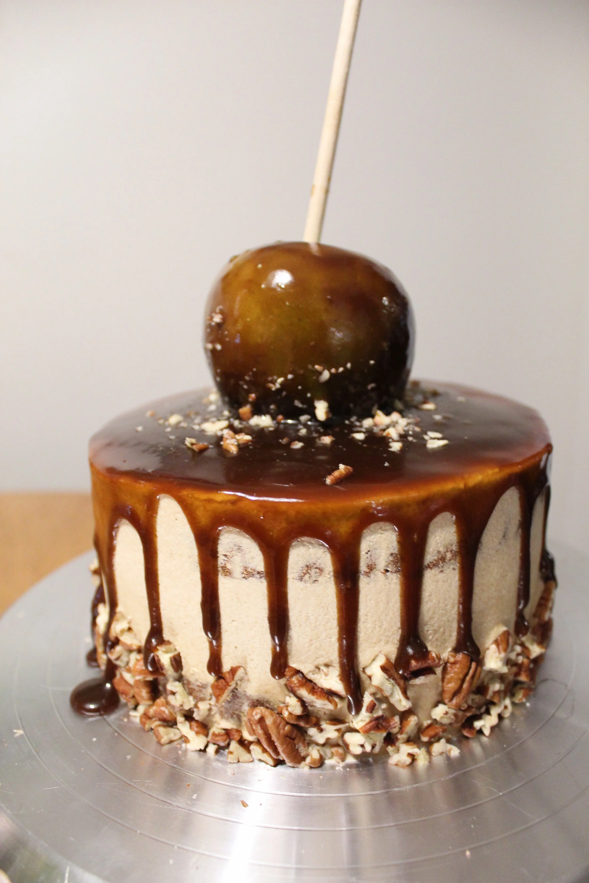 Round cake with beige frosting, chocolate glaze dripping from the top, chopped nuts around the base, topped with a caramel apple on a skewer.