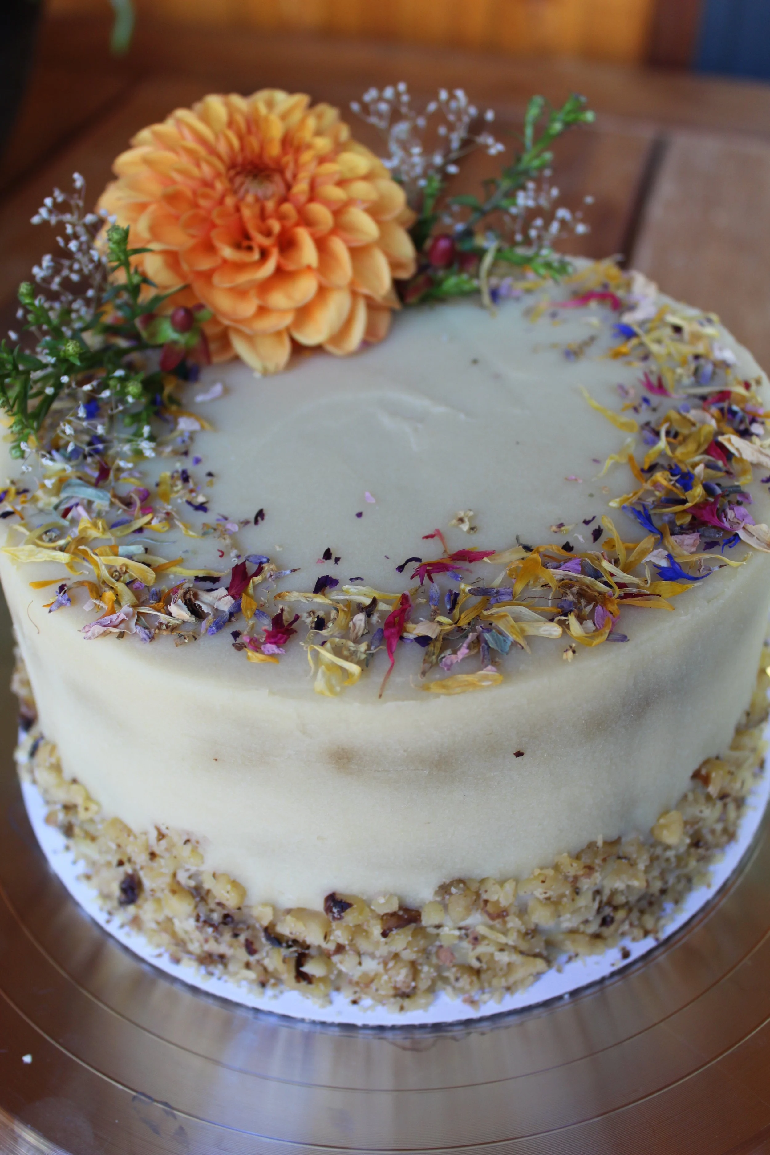 A layered cake decorated with a large orange flower, dried flower petals around the top edge, and chopped nuts around the bottom edge.