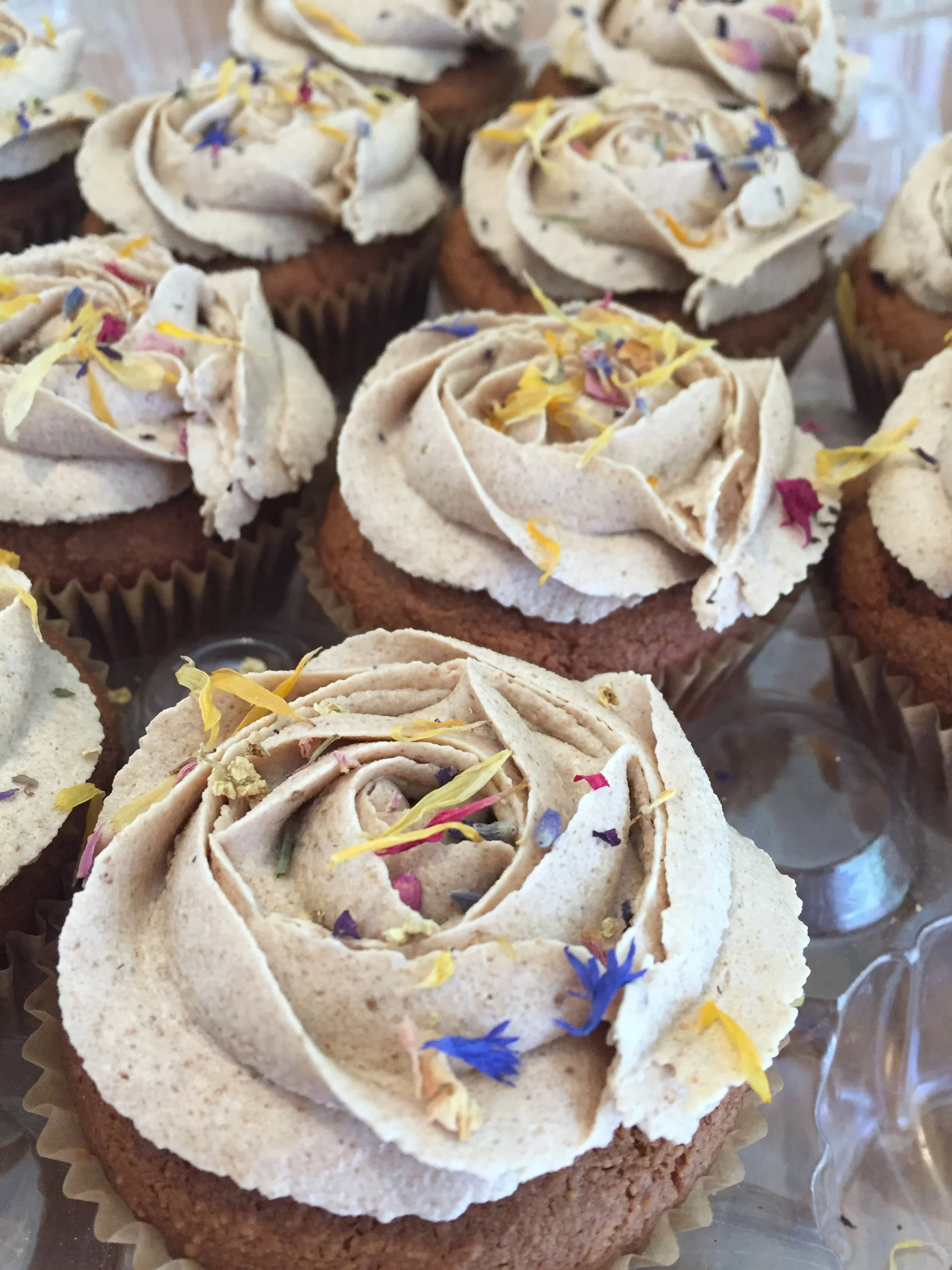 Mini cupcakes with chocolate bases topped with beige frosting and decorated with colorful edible flower petals.