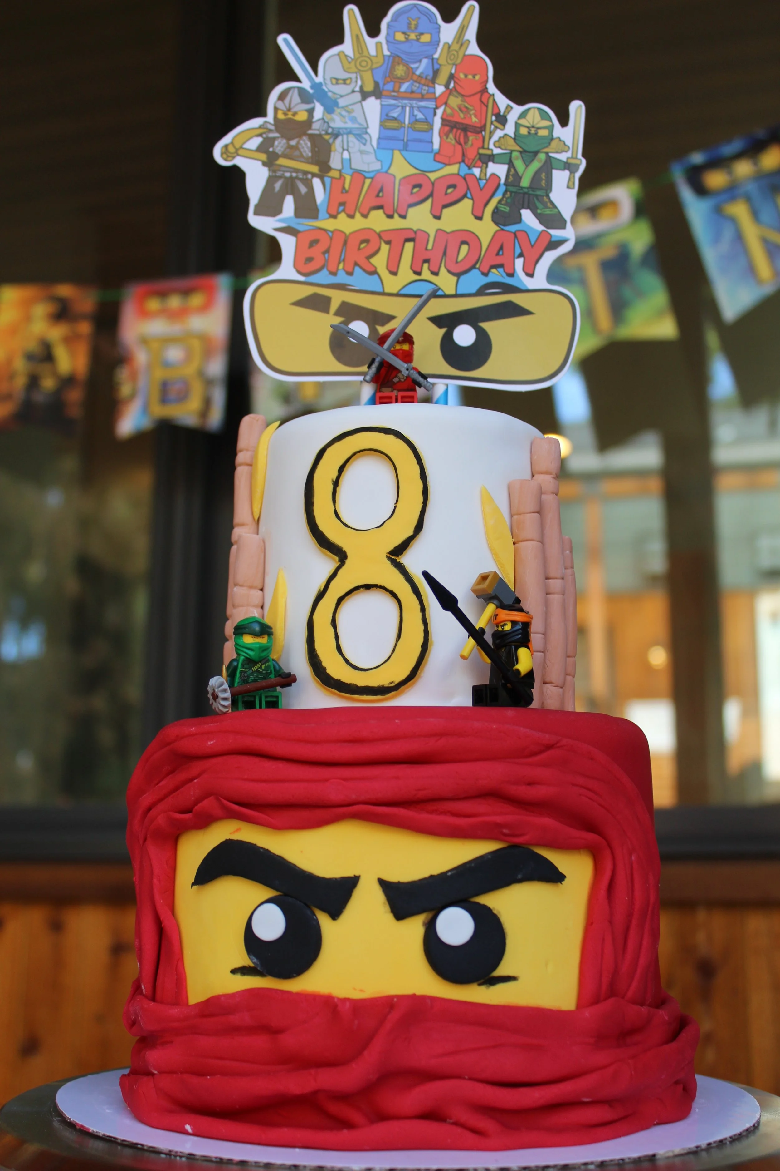 Lego Ninjago themed birthday cake with three tiers. The bottom tier resembles a Lego face with black eyebrows and eyes, wrapped in red fondant. The middle tier has a large yellow number 8 with a ninja figure standing beside it. The top tier features 