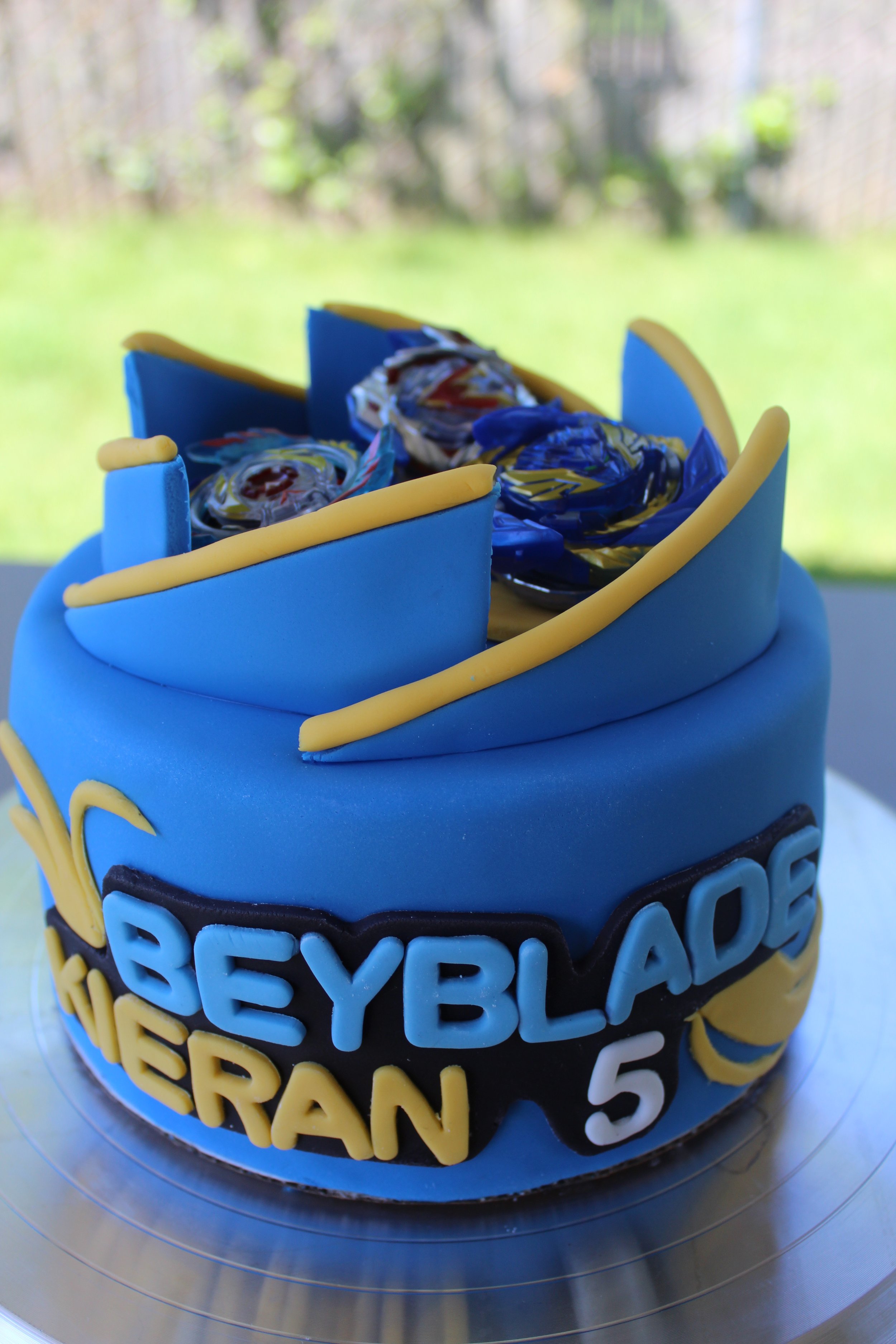 A birthday cake decorated in blue, yellow, and black colors with the words "Beyblades Kieferan 5" on the side, featuring Beyblade-themed decorations on top.