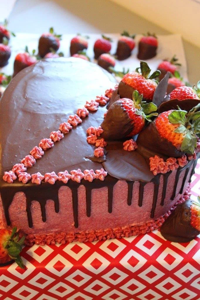 Chocolate-covered strawberry decorated heart-shaped cake with strawberries and pink decorative border.