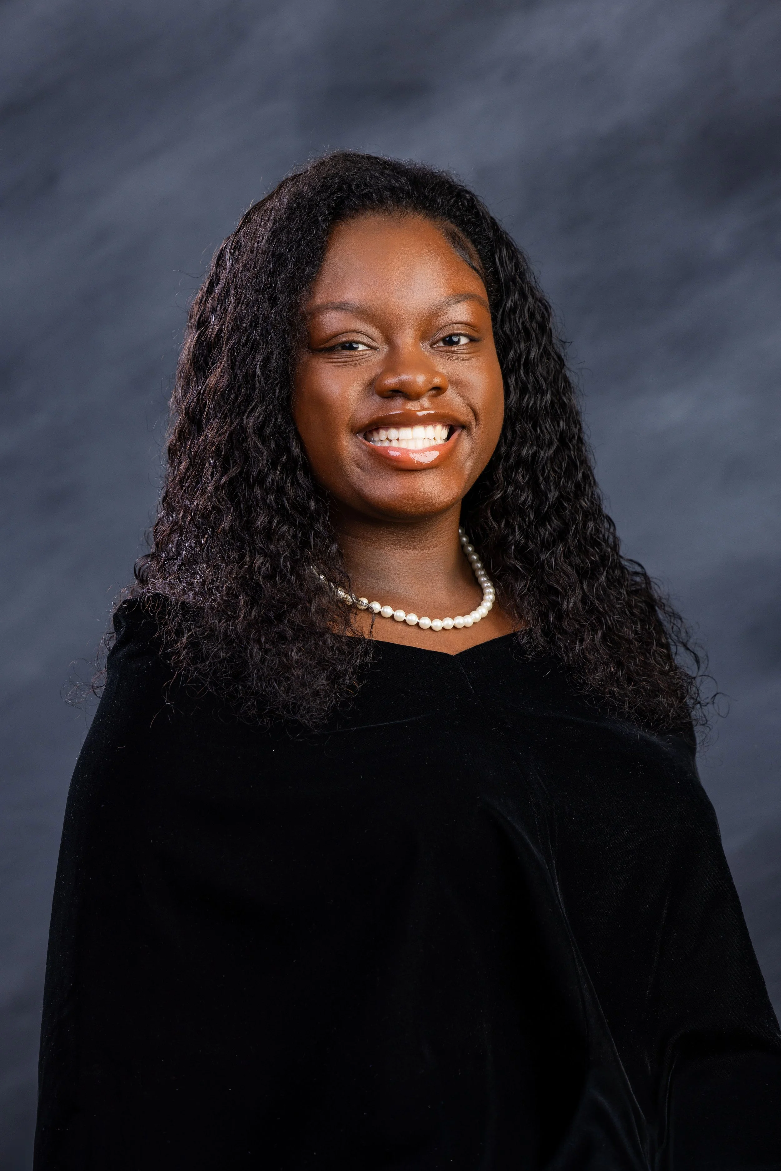 High school graduate girl with long wavy hair, wearing a black gown and a pearl necklace, smiling against a dark background.