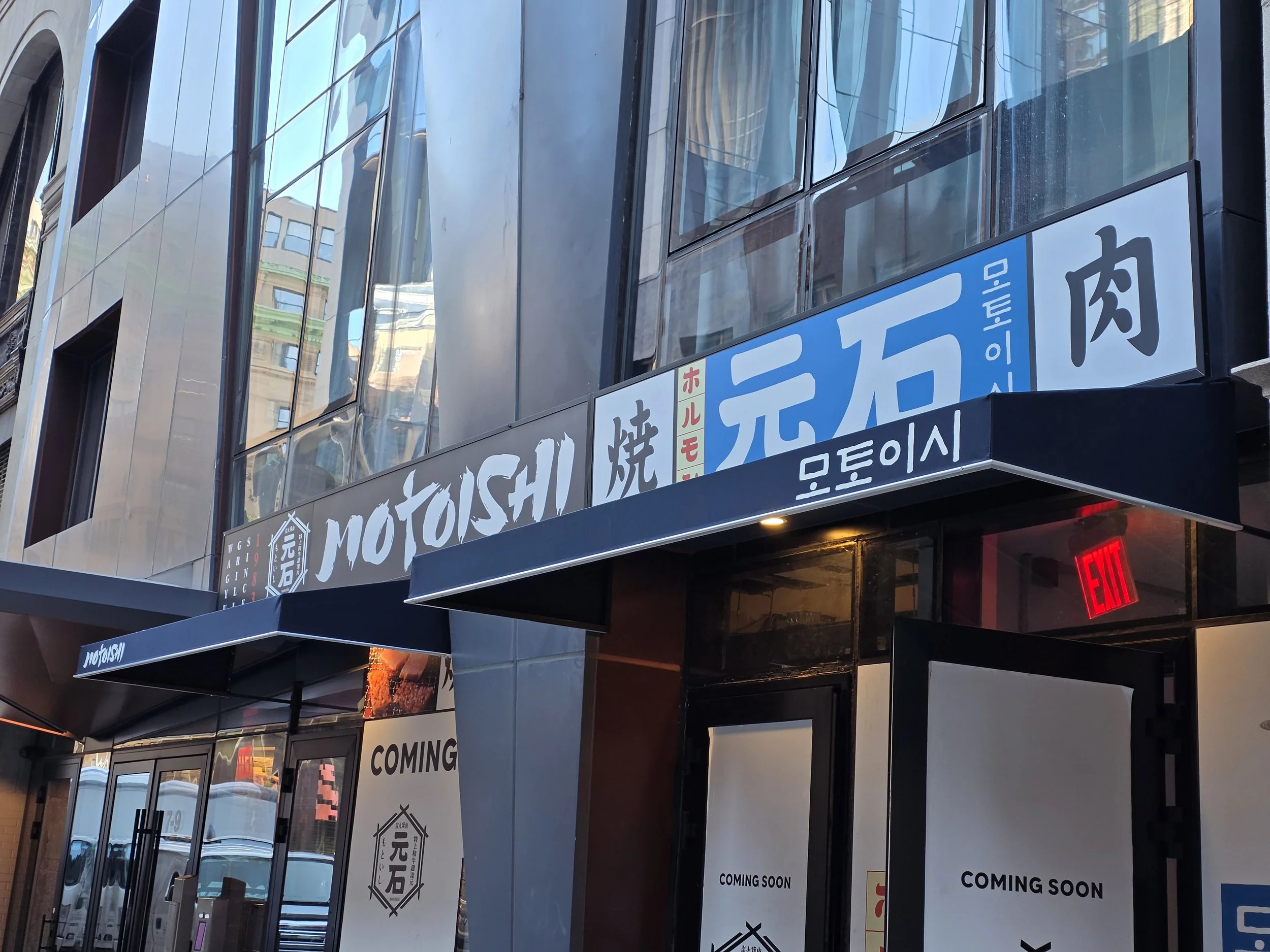 City street view with storefront signs, including one for a restaurant named Motoyoshi, with Korean signage and a "Coming Soon" poster, glass building reflections, and an illuminated exit sign.