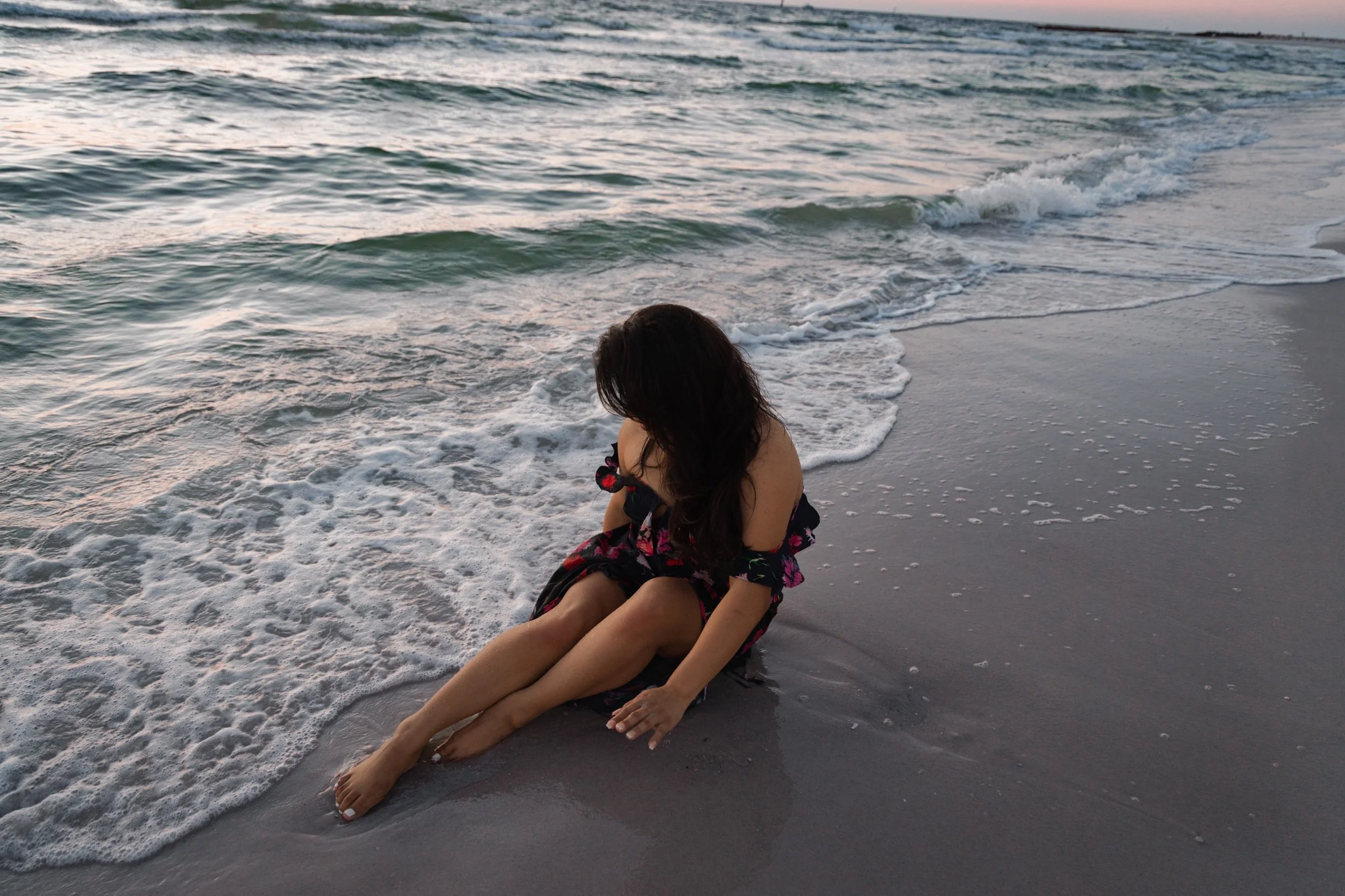 A woman in a floral dress sits on the shoreline as gentle ocean waves roll in, captured during a serene sunset. The soft light reflects off the water, creating a peaceful coastal scene with natural textures of sand, sea foam, and rippling waves.