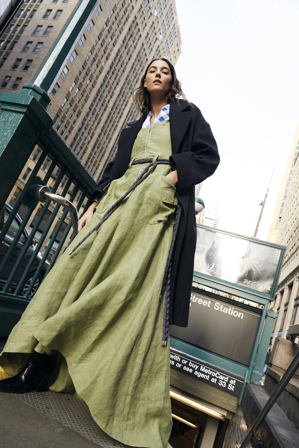 Fashion model wearing a long green dress and black coat standing near a subway entrance in a cityscape with tall buildings.