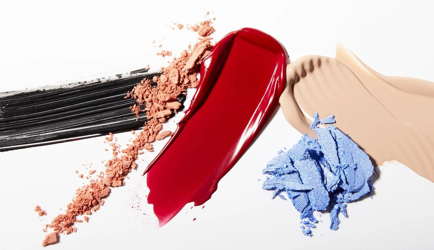 Smudges and smears of colorful makeup products on a white surface, including black mascara, pink and blue eyeshadow, red lipstick, and beige foundation.