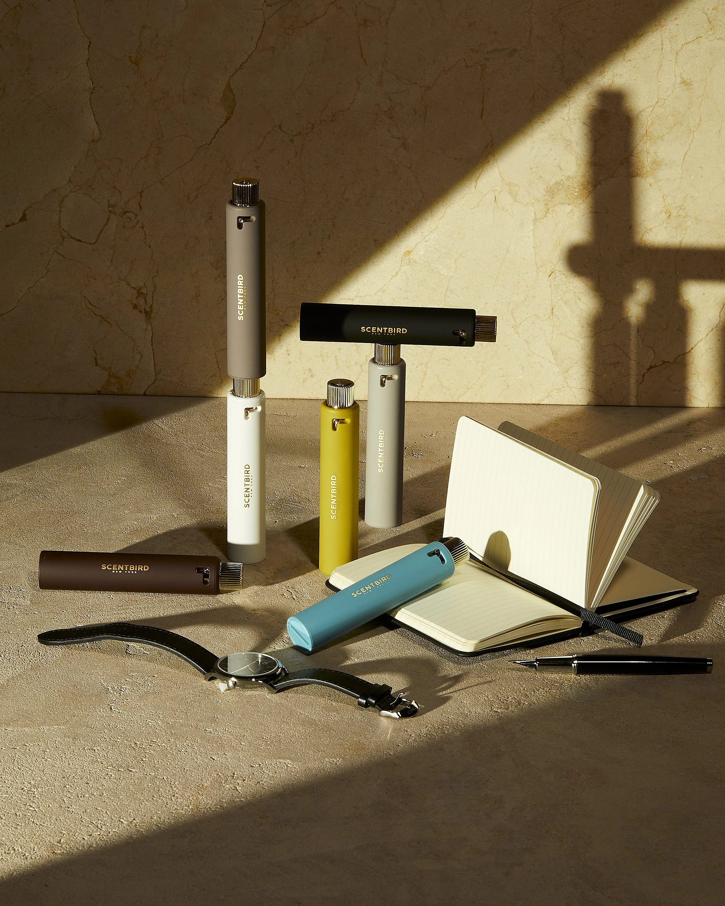 Assorted Scentbird perfume bottles placed on a surface alongside a wristwatch, an open notebook, and a pen, with a shadow cast on the background wall.