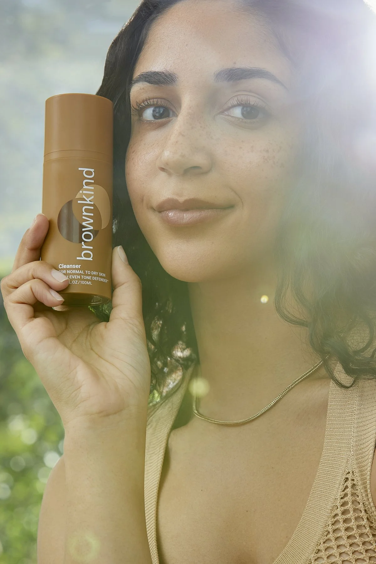 Beauty photography of a smiling model holding a brown glass skincare bottle outdoors in natural light, showcasing a sustainable skincare product for a beauty brand campaign by Adrianna Favero, NYC beauty photographer and director.