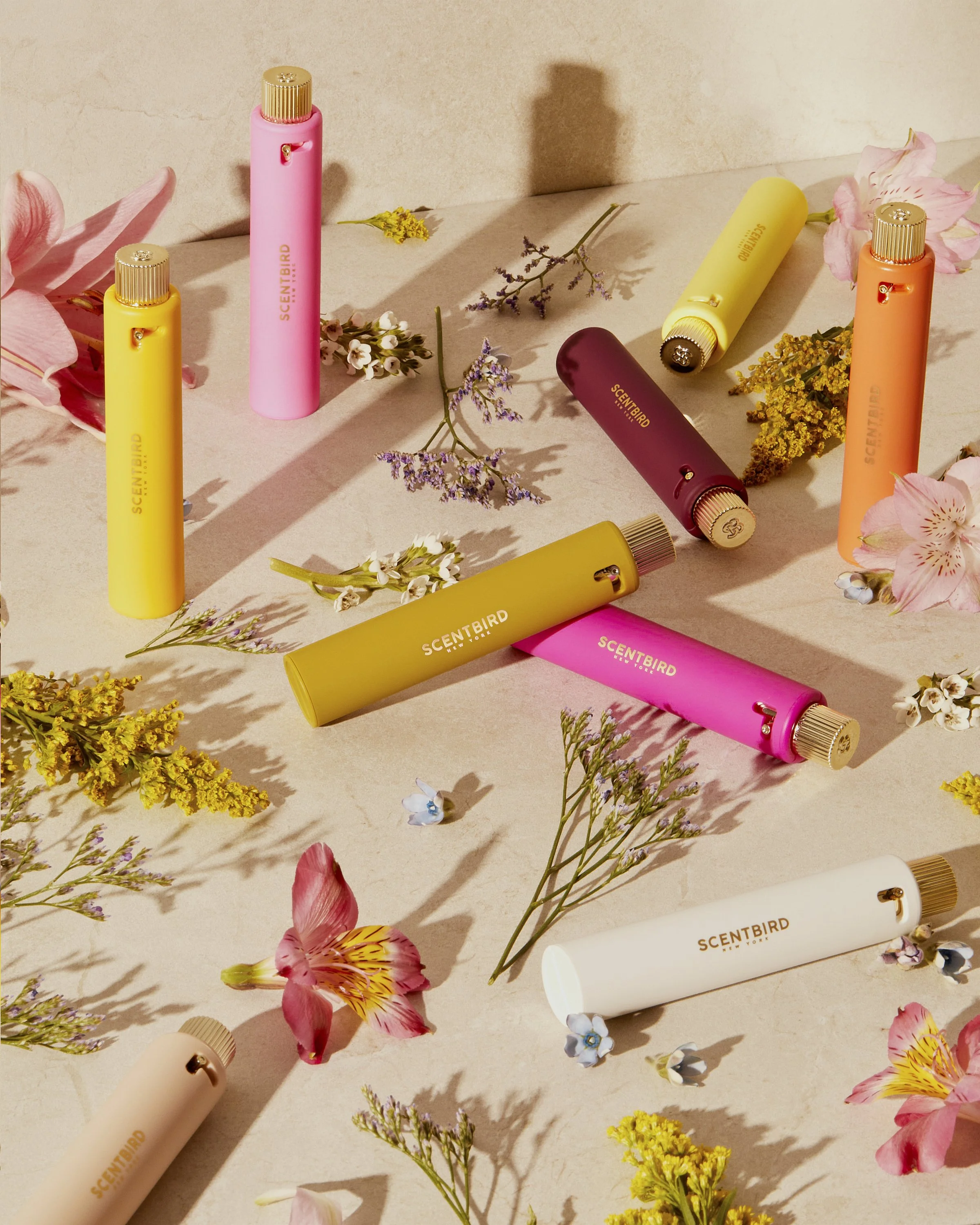 Colorful Scentbird perfume containers surrounded by assorted flowers on a light surface.