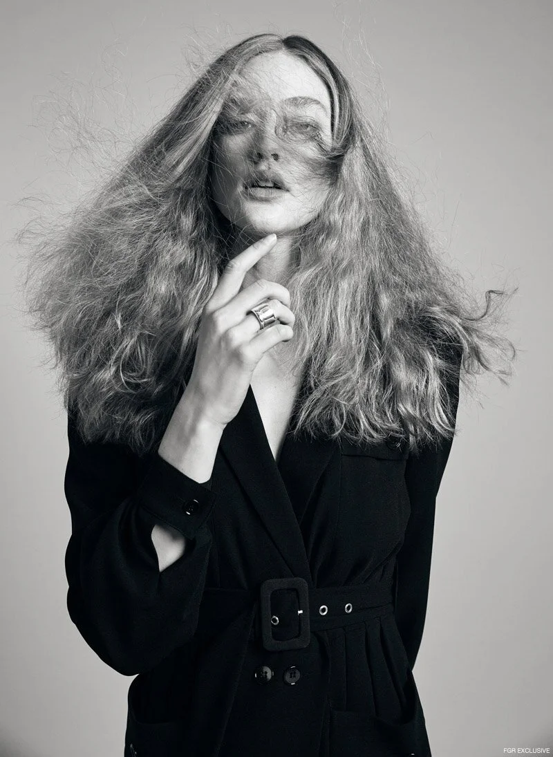 Black and white photo of a woman with long, voluminous hair wearing a black outfit and rings, posing with her hand near her chin.