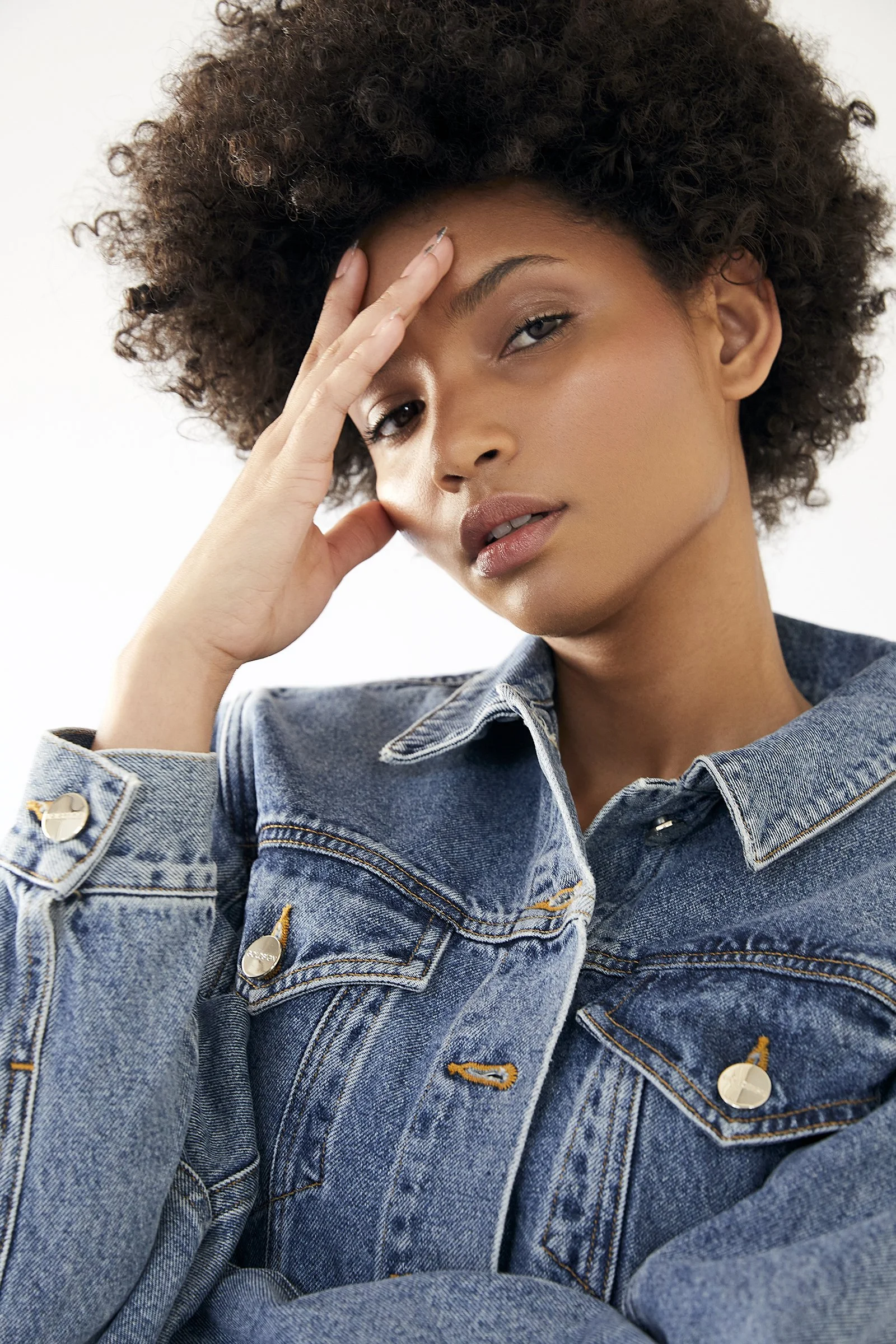 Person wearing a denim jacket with an afro hairstyle, hand resting on head.