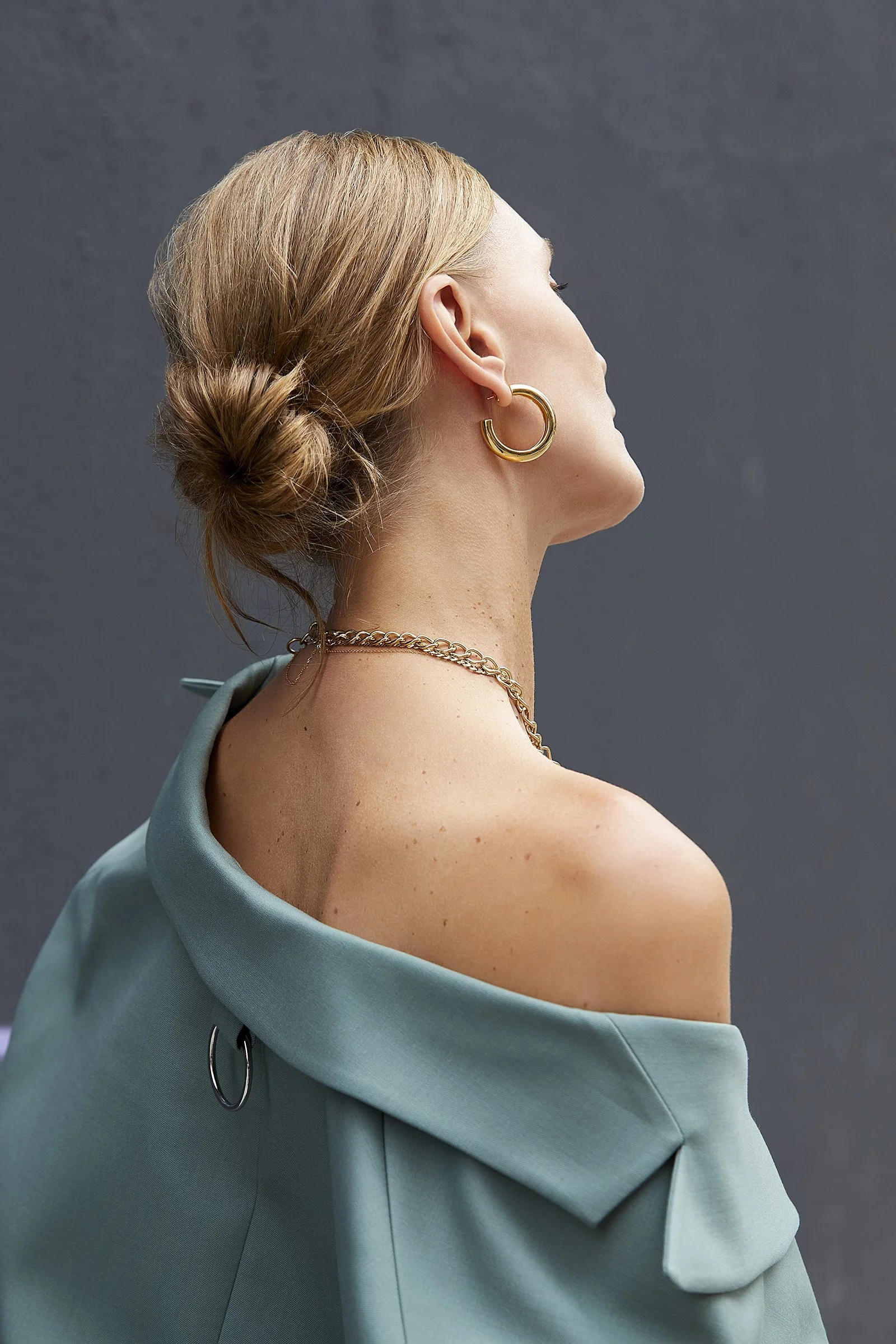 Blonde model with her hair in a bun, wearing an off-shoulder teal top and a delicate gold necklace, shown in profile, for an Aurate fall jewelry collection editorial.