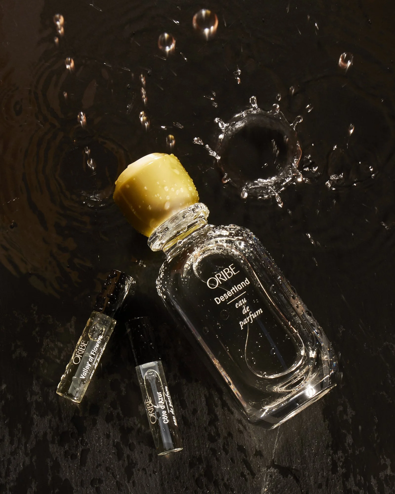 Oribe Desertland perfume bottle and two vials with water splashes on a dark background.