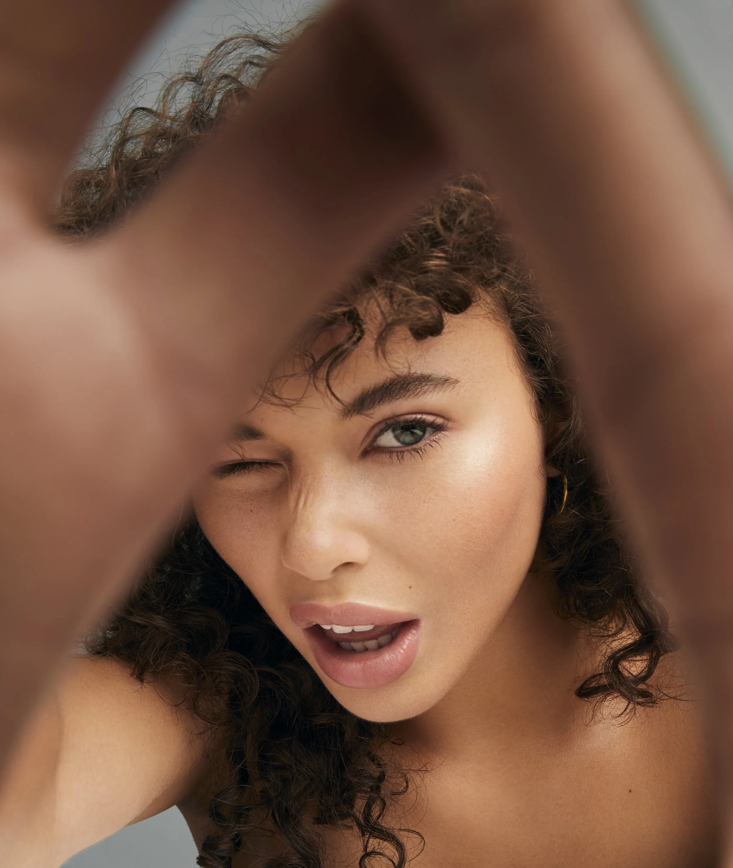 Woman with curly hair posing, winking and making a gesture with her hands in front of her face.