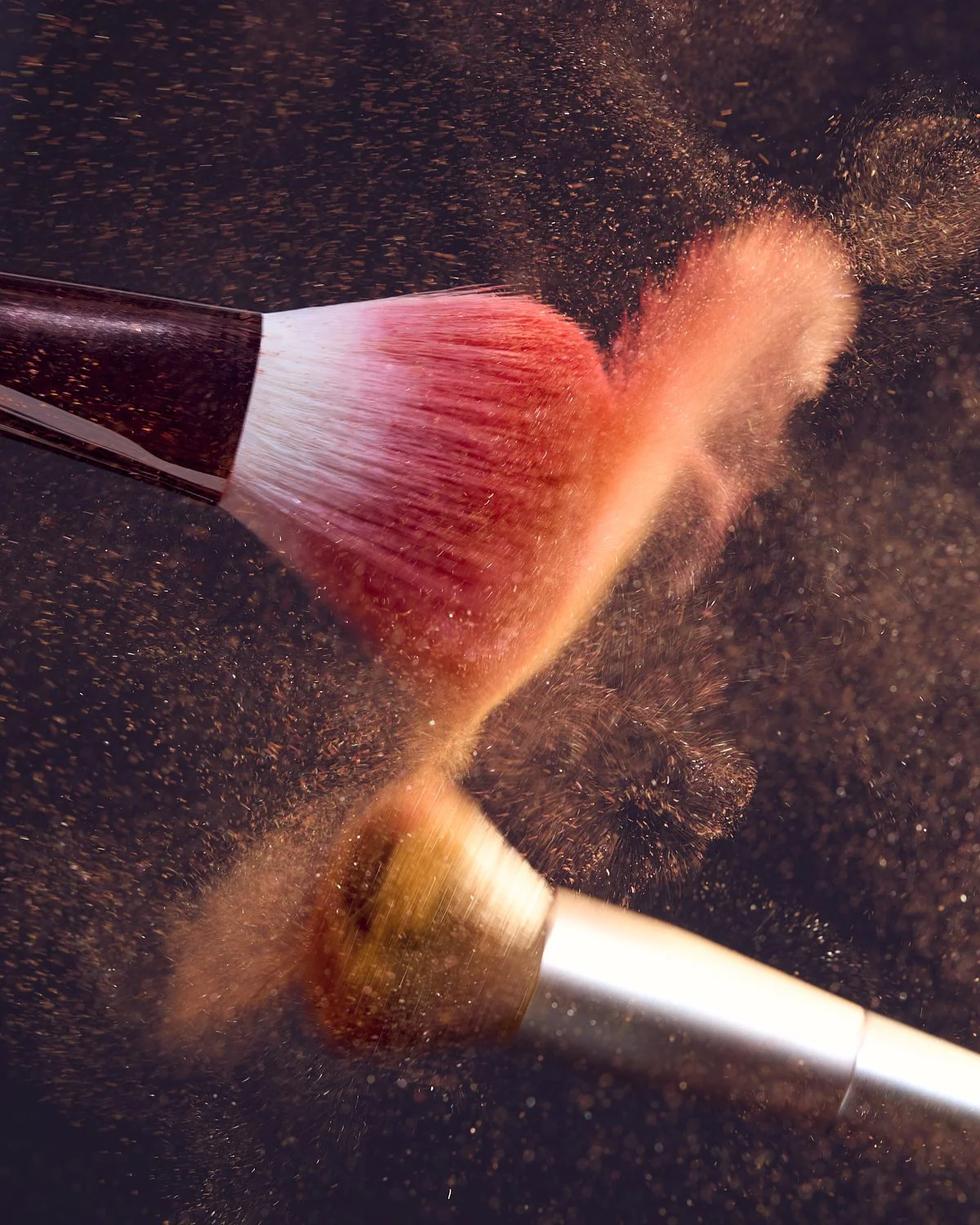 Dynamic motion shot of a makeup brush releasing a burst of coral pigment powder against a dark background, by Adrianna Favero.