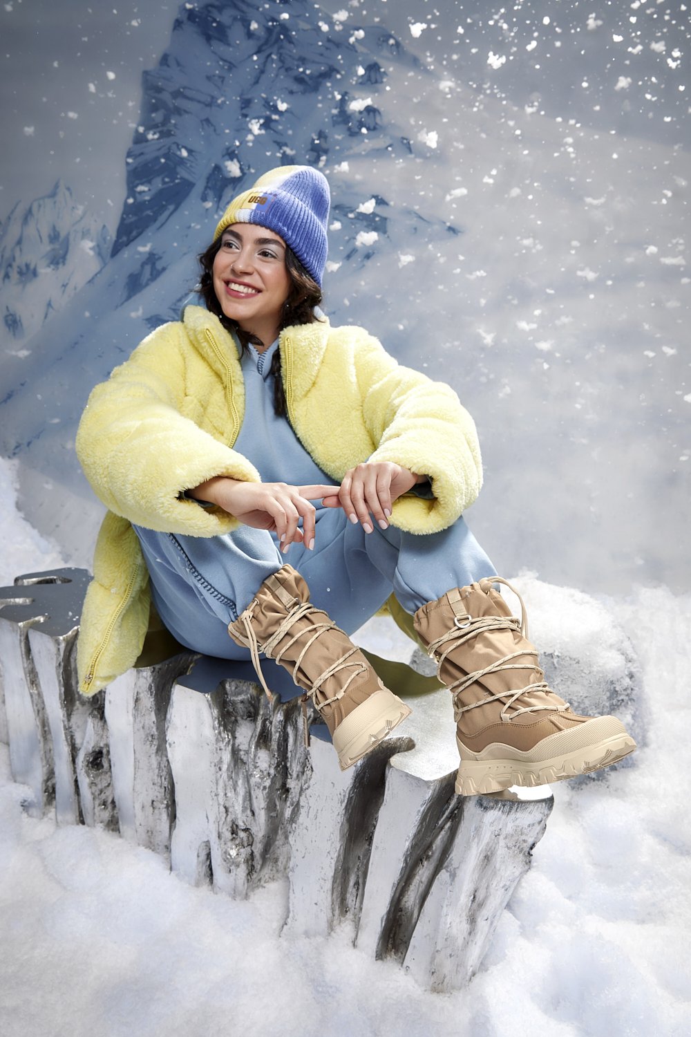 A smiling person wearing a yellow fur coat, blue knit hat, light blue pants, and tan snow boots sitting on a snowy log with a snowy mountain backdrop.