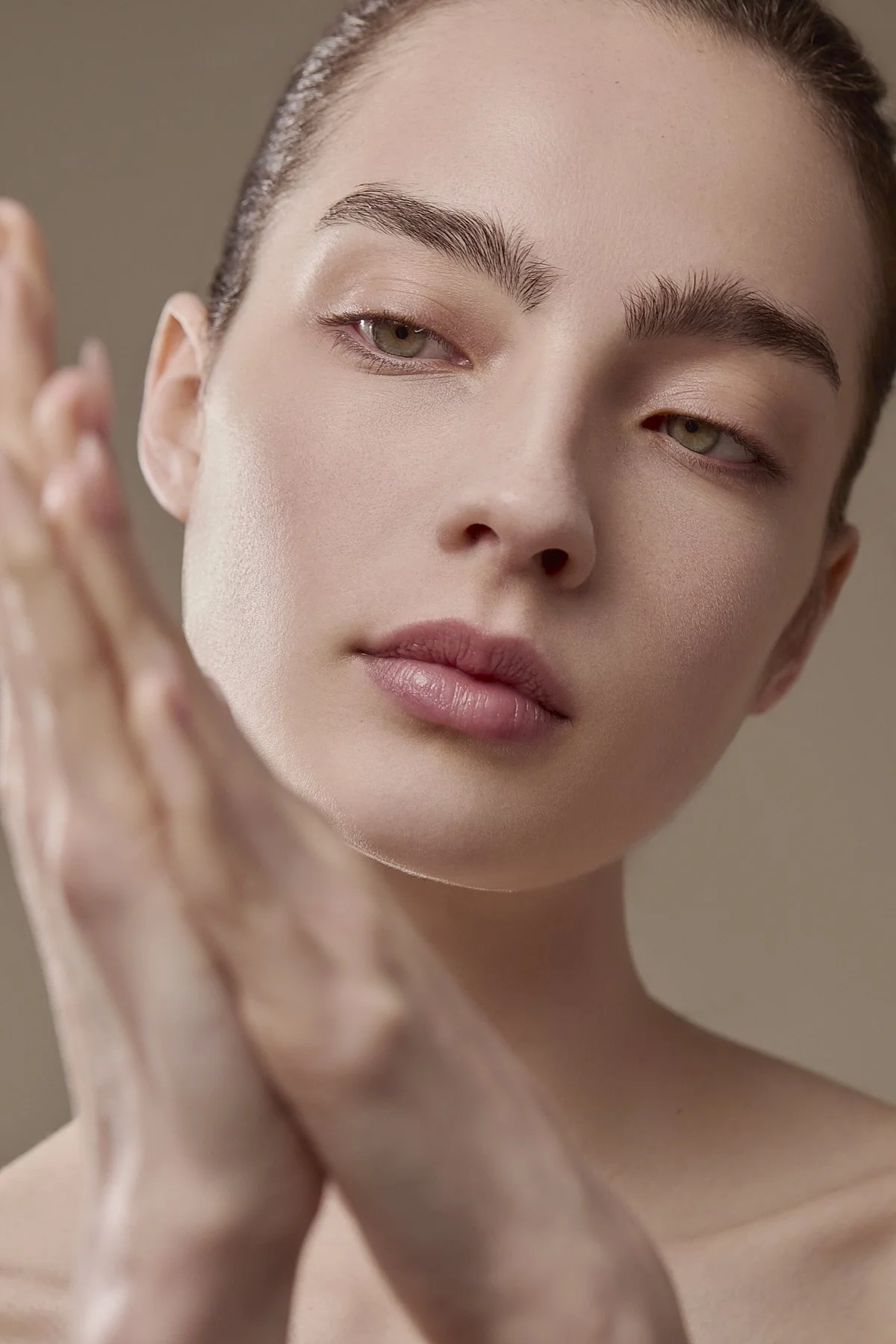 Soft beauty close-up of a model with hands gently framing her face and eyes closed, showcasing natural, glowing skin in warm neutral tones, commercial beauty photography by Adrianna Favero, NYC beauty photographer and director.