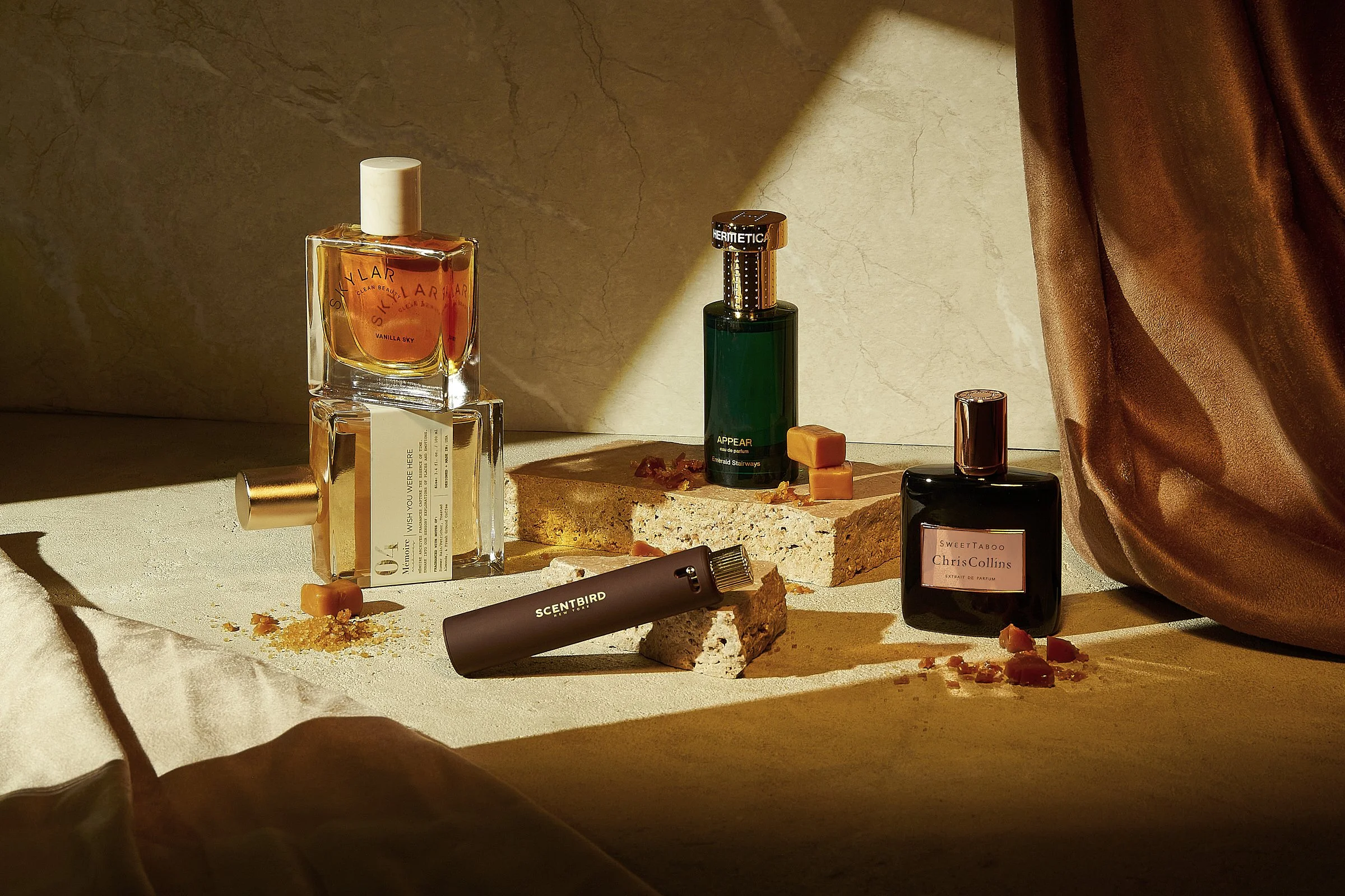 Various perfume bottles and a cylindrical scent holder on a textured surface with draped fabric in the background, surrounded by caramel pieces.