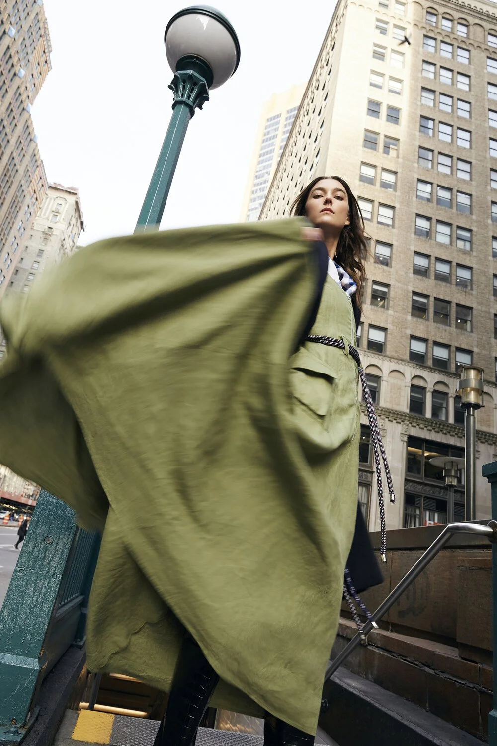 A woman in a flowing green coat standing near a city street entrance, surrounded by tall buildings and a lamppost.