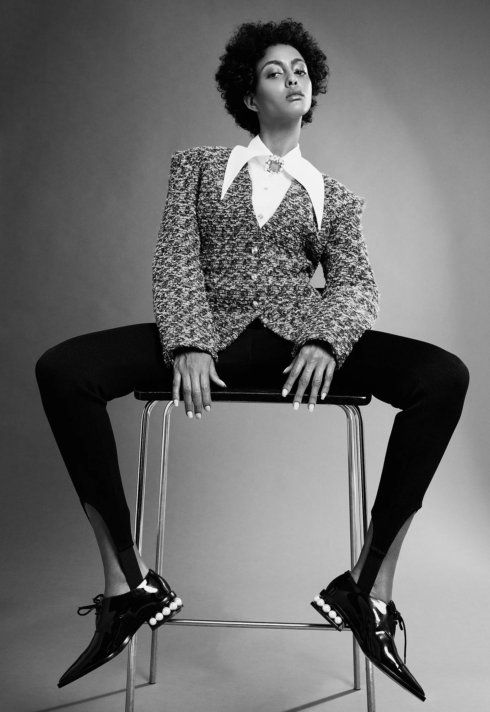 Person sitting on a stool wearing a textured jacket, white shirt with large collar, black pants, and shiny shoes with pearl accents.