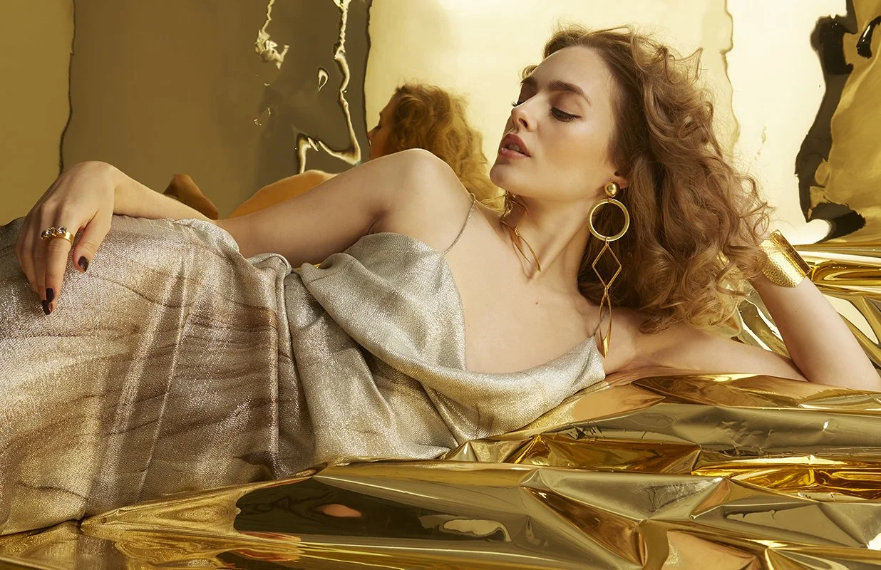 FAJO Magazine holiday fashion editorial: model reclining on a crinkled gold foil backdrop in a metallic gold slip dress, wearing statement drop earrings and gold rings, photographed by Adrianna Favero.