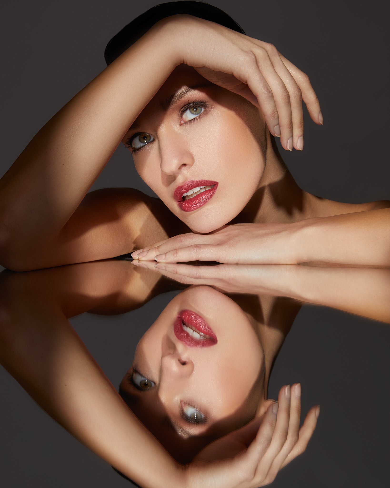 A woman posing with her hands framing her face, reflected in a mirror, showcasing her makeup and facial features.