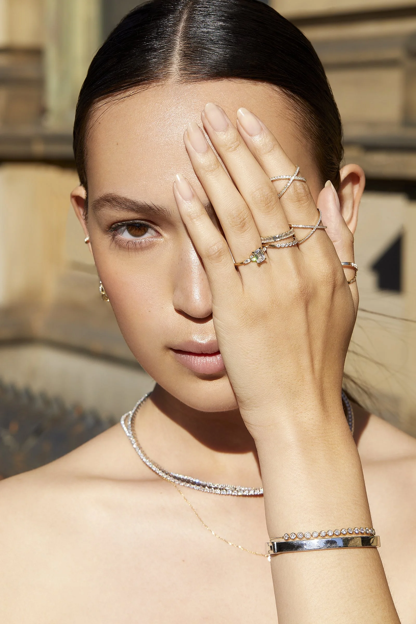 Model shading her face with her hand, showing a delicate gold bracelet and stacked rings, sleek dark hair and soft neutral background, for an Aurate New York jewelry editorial.
