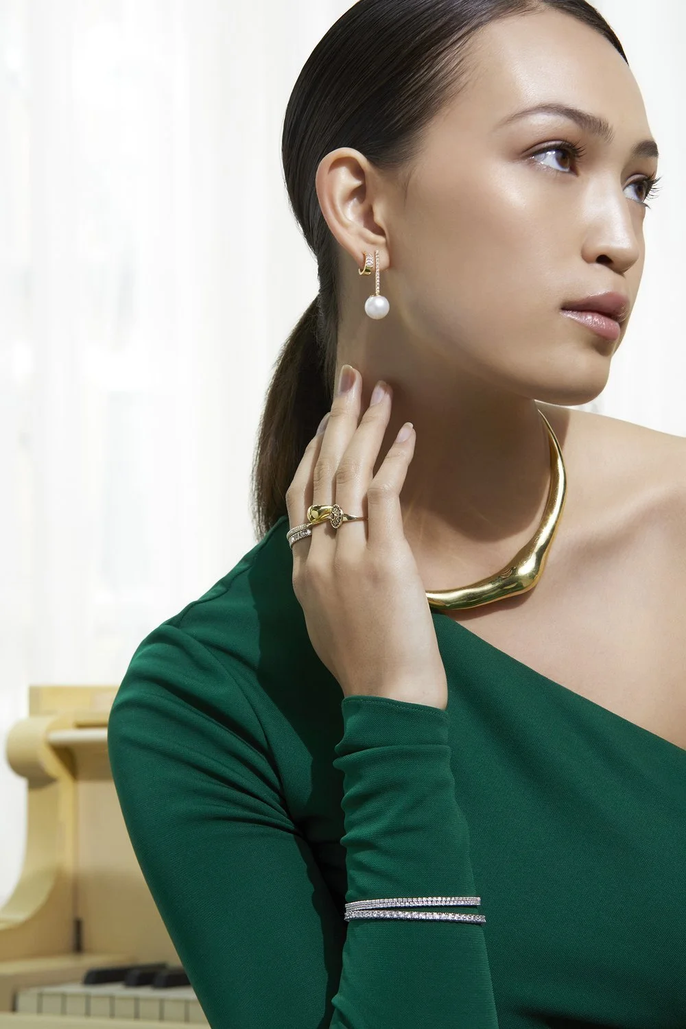 Woman in a green dress wearing gold jewelry, including a necklace, ring, and pearl earring, posing with hand near her face.