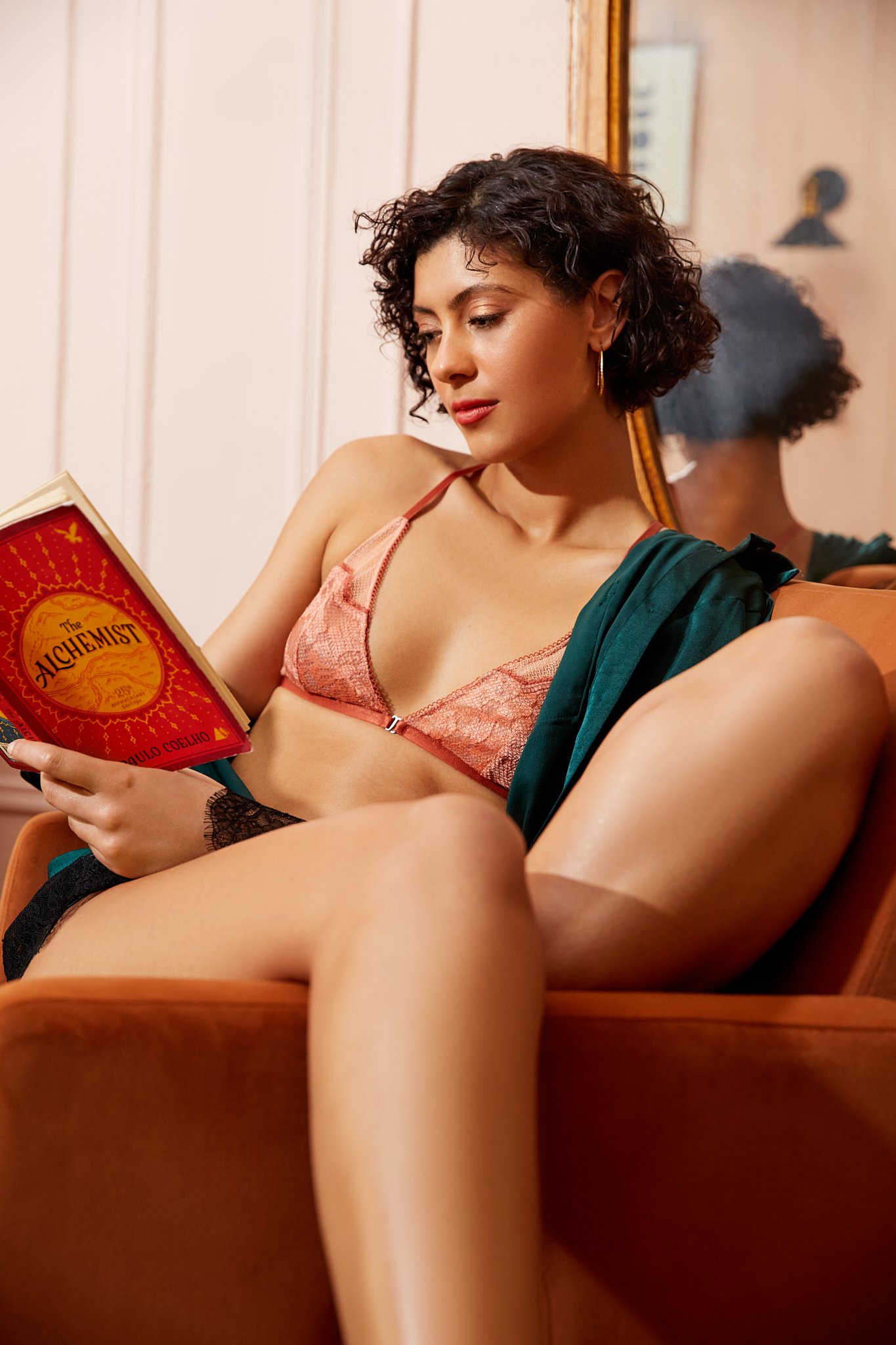 Person reading "The Alchemist" by Paulo Coelho, seated in an orange chair, wearing a coral-colored bralette.