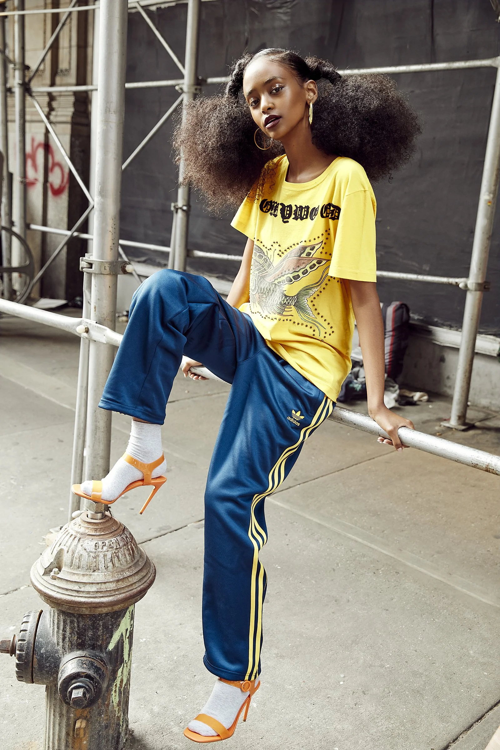 Person in a yellow graphic t-shirt, blue Adidas track pants with yellow stripes, wearing orange high heels with white socks, sitting on a metal railing and leaning against a fire hydrant on a city street.