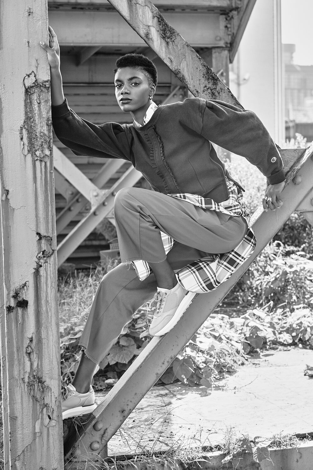 Fashionable person posed on industrial beams, wearing a jacket and plaid shirt.
