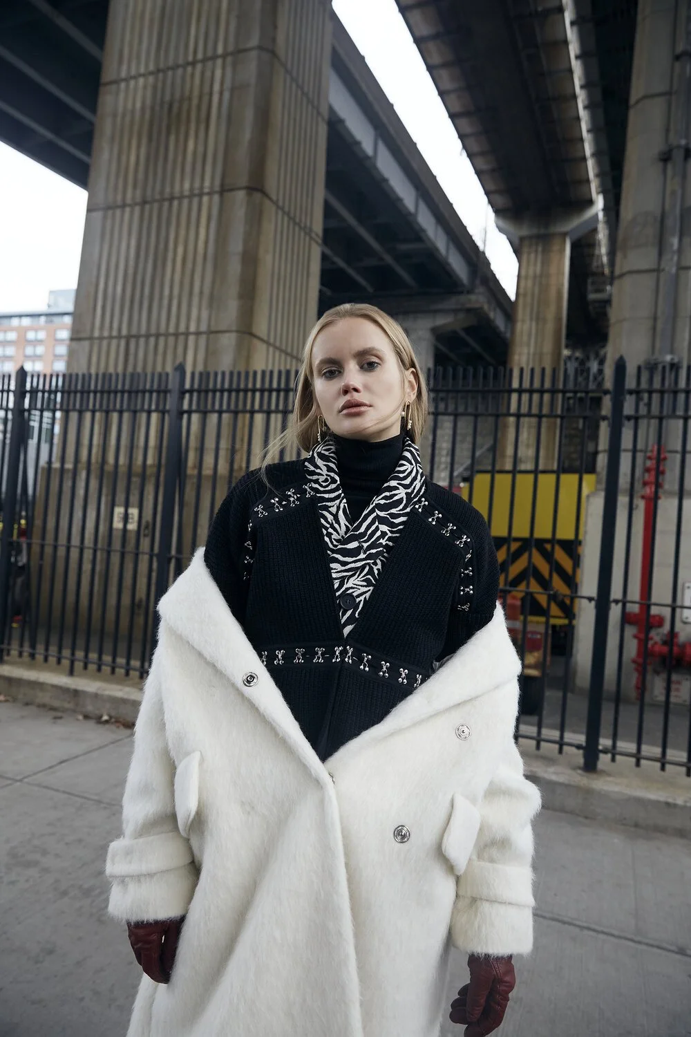 Person wearing a white coat and patterned scarf standing under an overpass with a fence in the background.