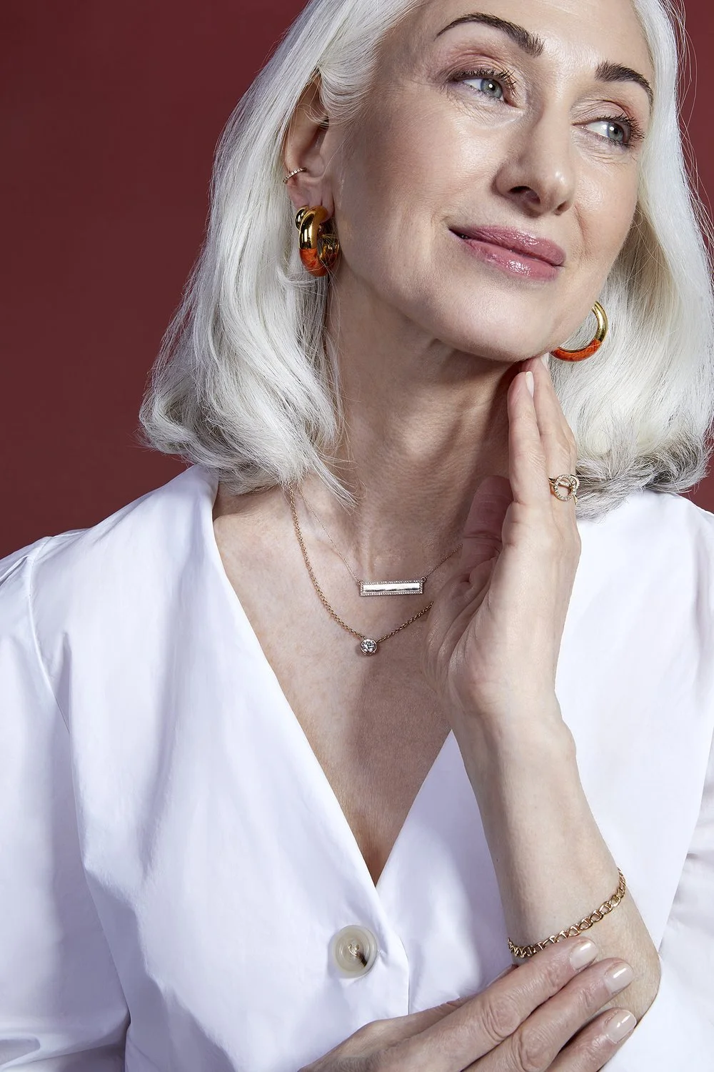 Woman with silver hair wearing gold hoop earrings, gold necklaces, a ring, and a bracelet, dressed in a white top against a red background.