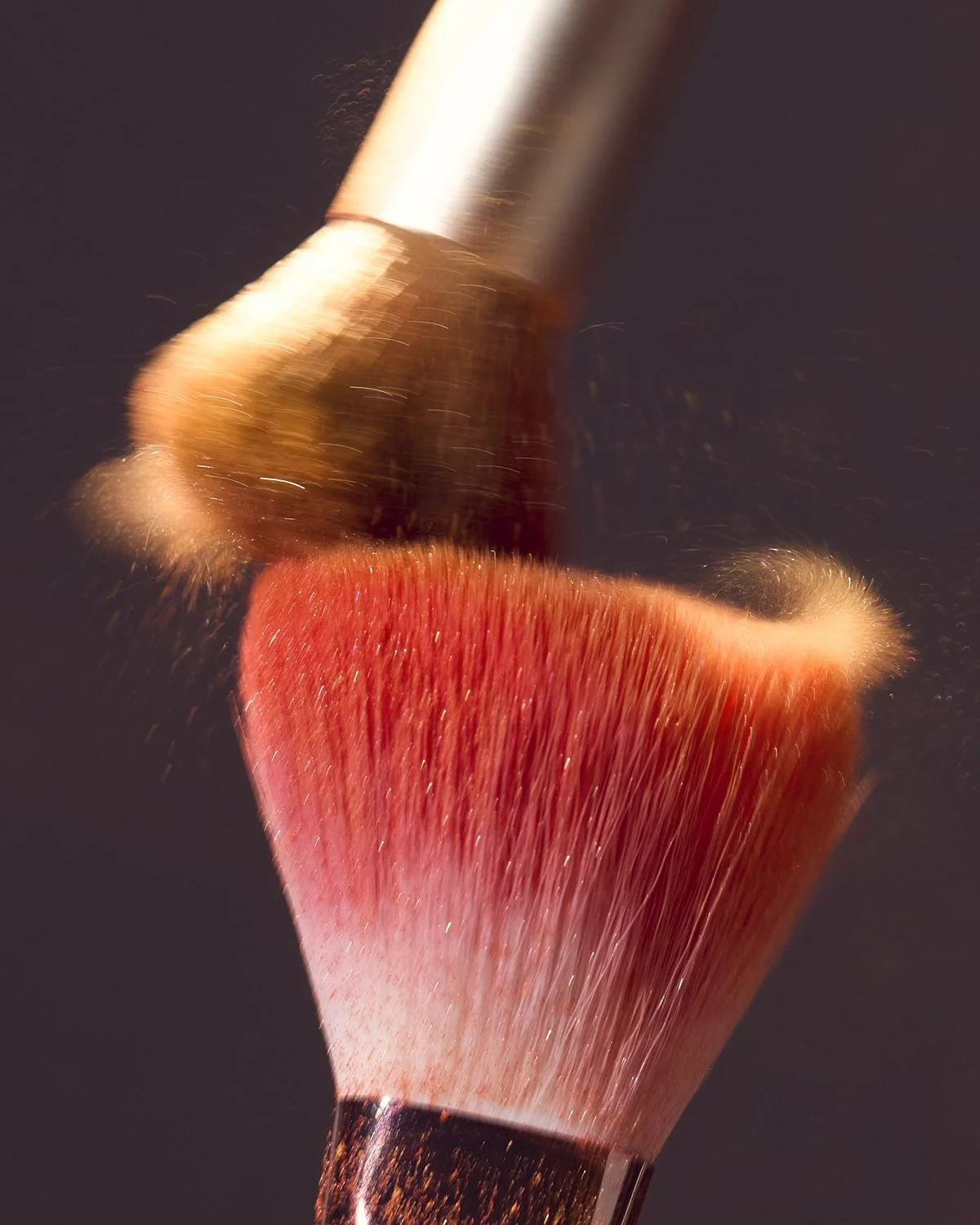 Close-up of a fluffy makeup brush coated in coral pigment powder against a dark background, by Adrianna Favero.
