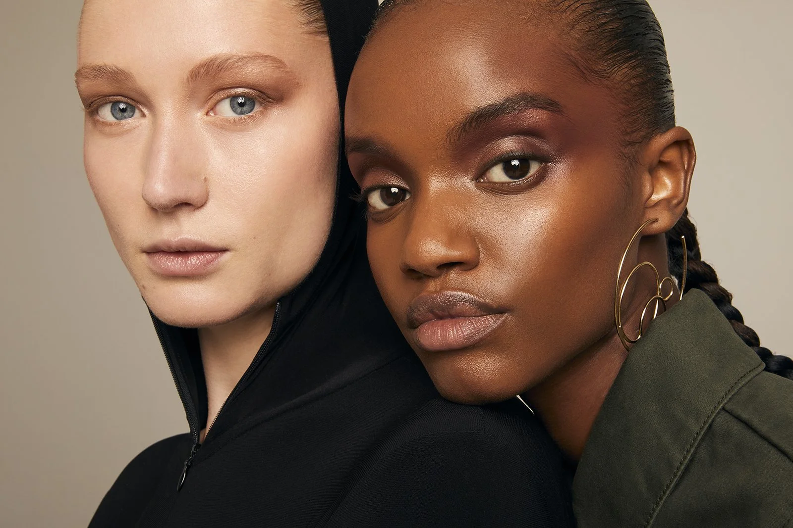 Two women posing together, close-up, with natural makeup and different skin tones. One is wearing a black top, the other in an olive green top with gold hoop earrings.