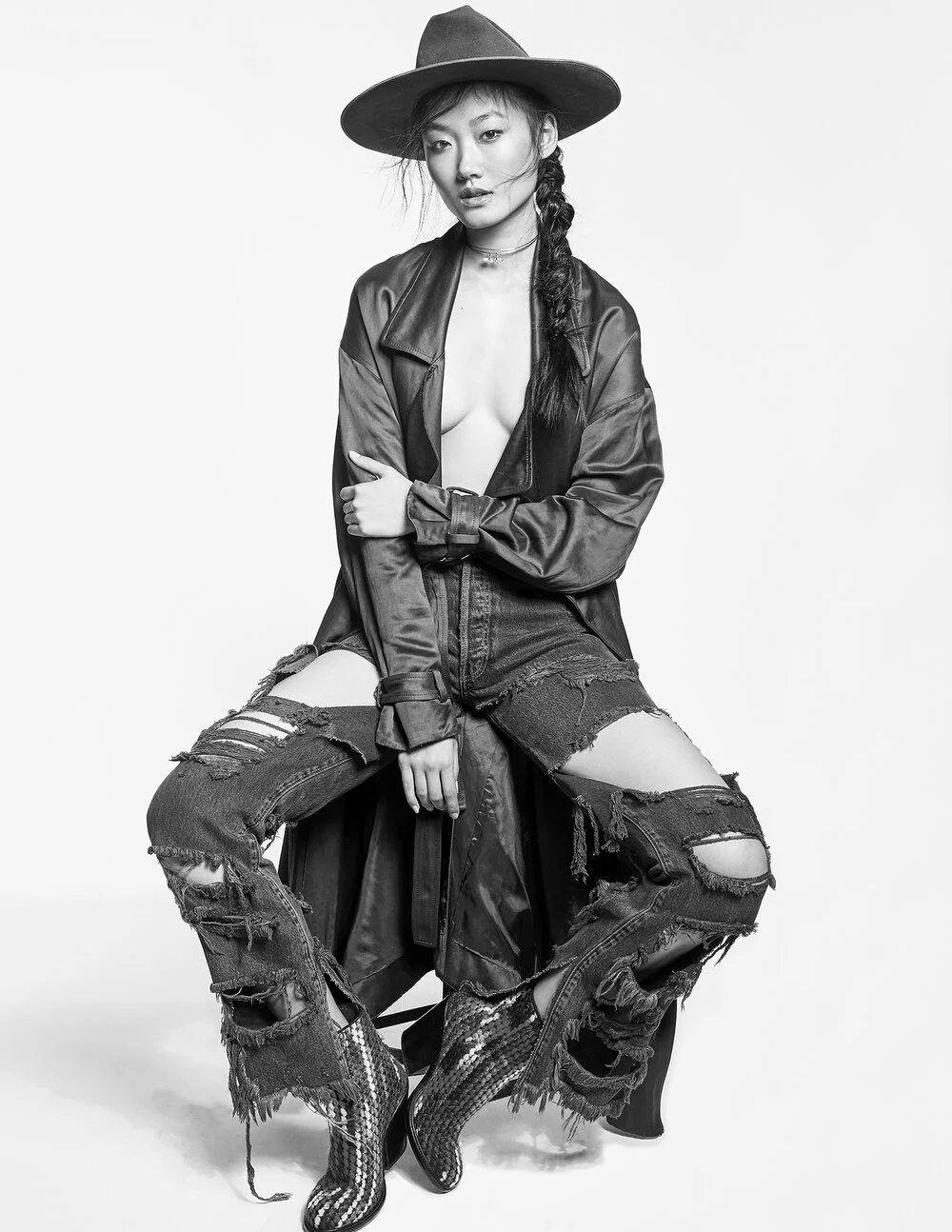 Fashion model sitting, wearing a wide-brimmed hat, long coat, and ripped jeans, with knee-length boots.