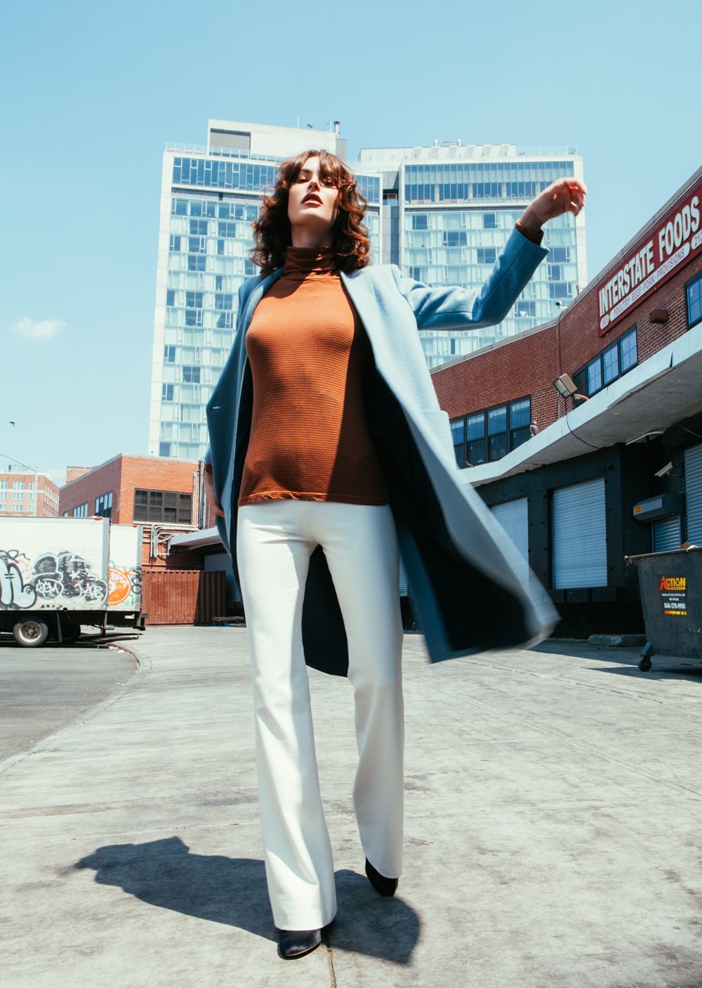 Fashionable woman in urban setting wearing a blue coat, brown top, and white pants.