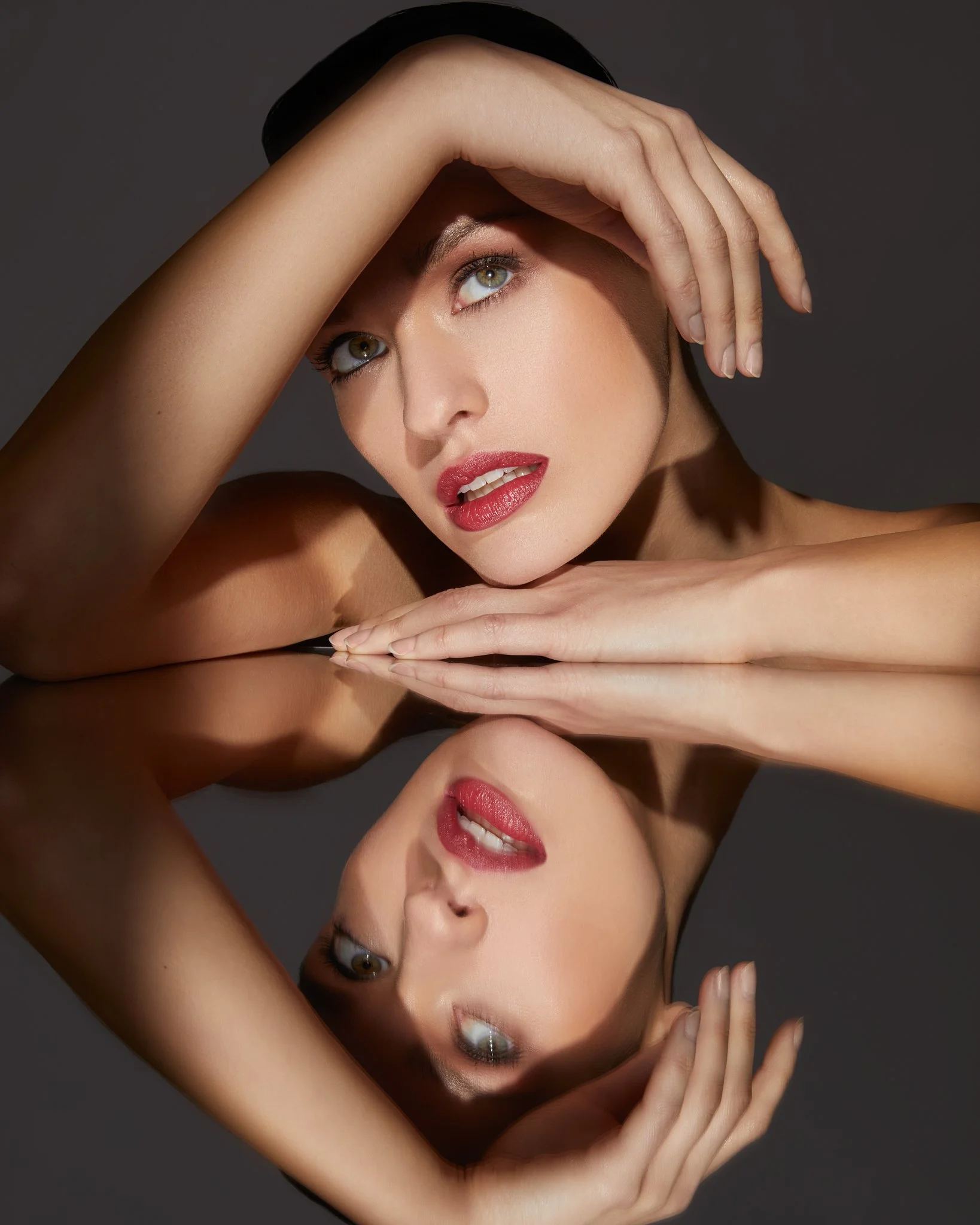A woman with styled makeup and red lipstick looking into a reflective surface, which shows her reflection. She has her hands placed dramatically around her face.