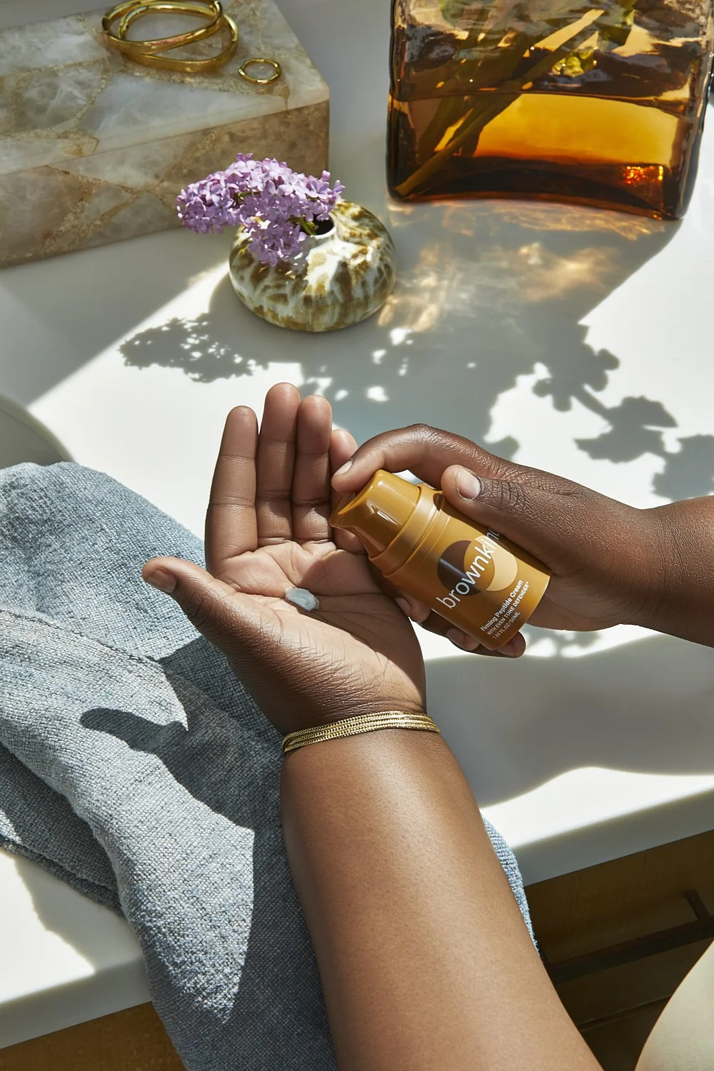 Hands holding a small amber product bottle beside dried flowers on a marble table, by Adrianna Favero.