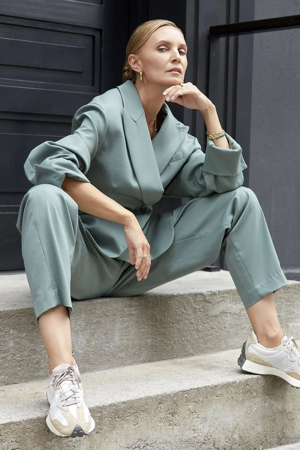 Woman in a green suit sitting on concrete steps with a contemplative expression, wearing white sneakers and gold hoop earrings.