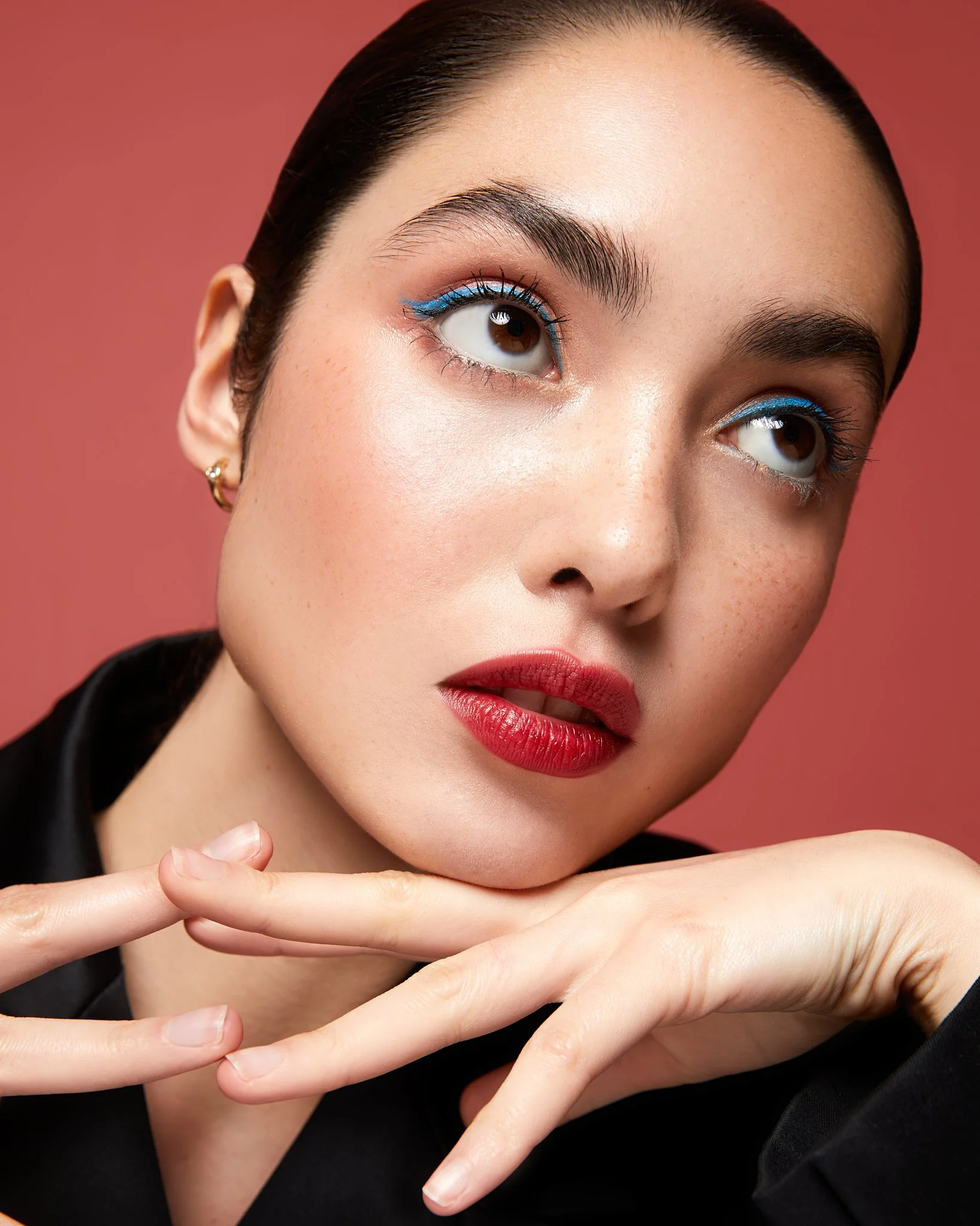 Close-up of a woman with dark hair, wearing red lipstick and blue eyeliner, posing with hands resting on her chin against a red background.