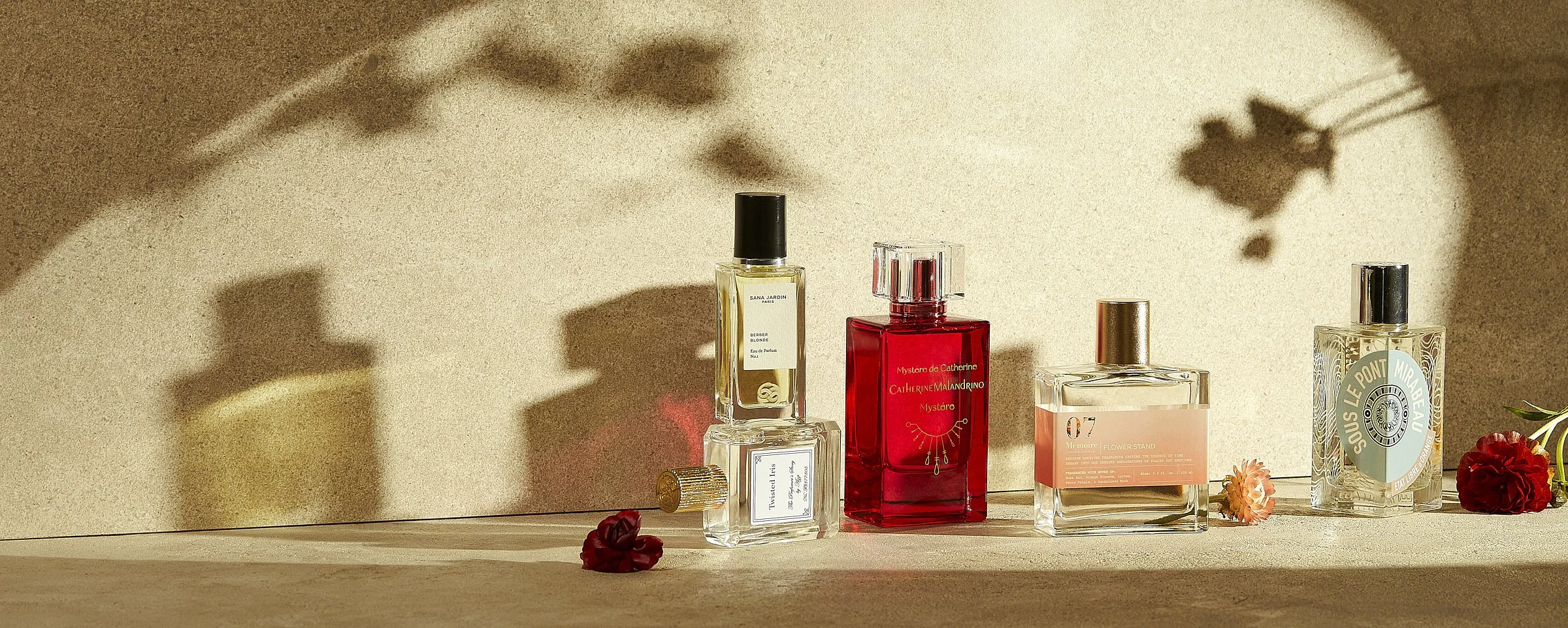 A display of various perfume bottles arranged on a textured surface with soft shadows and scattered flowers. The scene features five distinct bottles of different shapes and colors, including a prominent red bottle at the center. The setting is warml