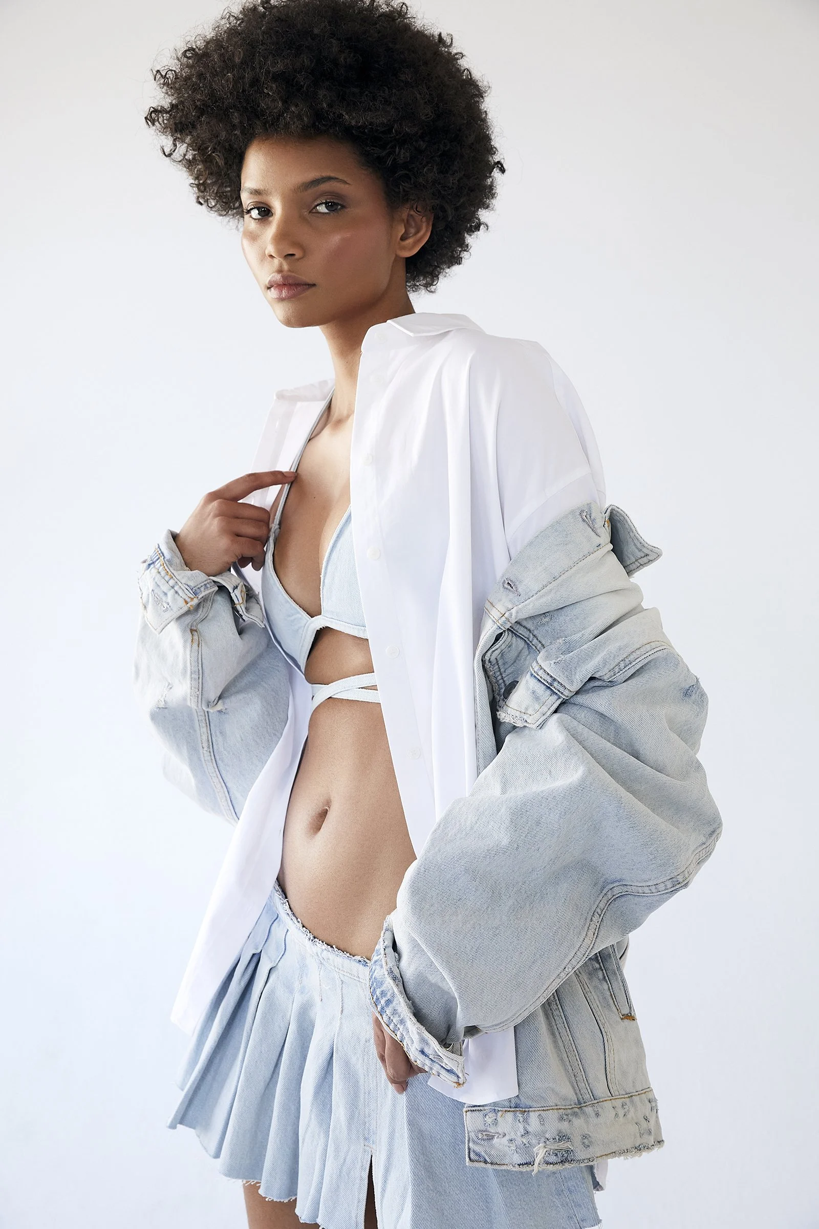 Woman wearing a denim bralette, skirt, and jacket with a white shirt underneath, looking at the camera in a minimalist setting.
