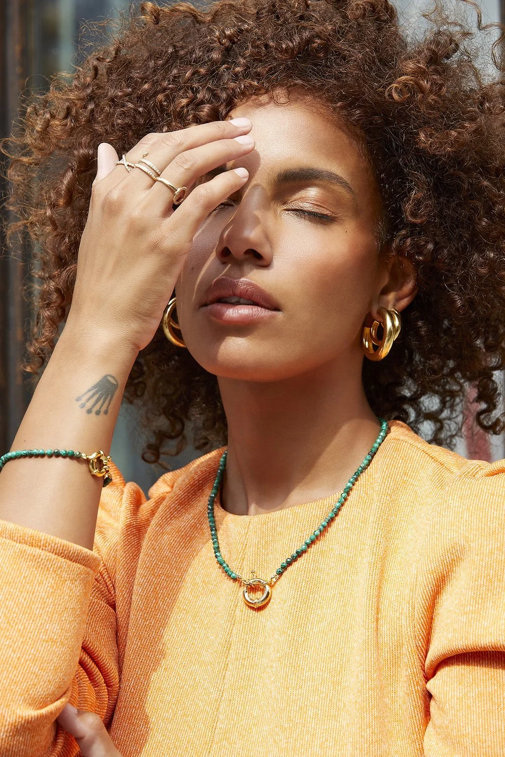 Woman with curly hair wearing large gold hoop earrings, an orange top, and green beaded jewelry.