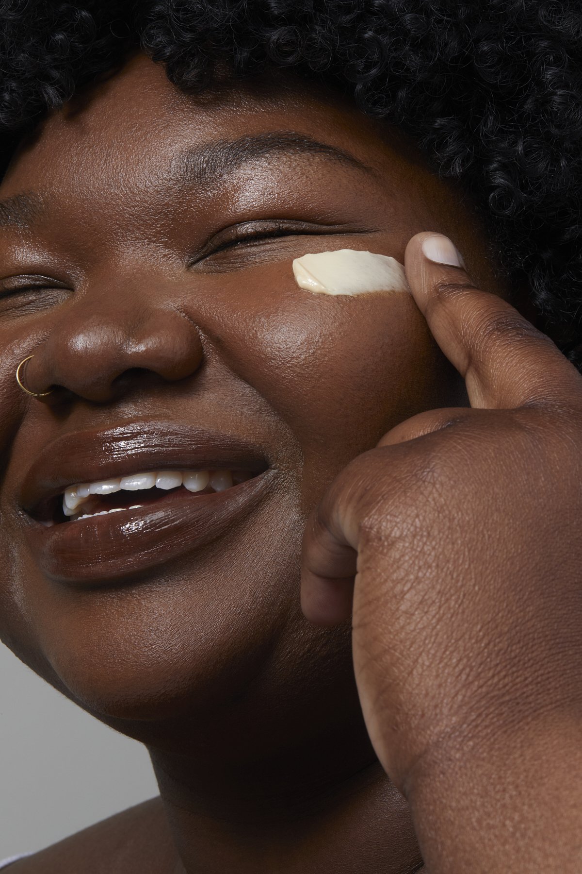 Beauty close-up of a smiling model with deep, radiant skin applying a dab of glow cream to her cheek, showcasing luminous skincare results for a beauty brand campaign by Adrianna Favero, NYC beauty photographer and director.